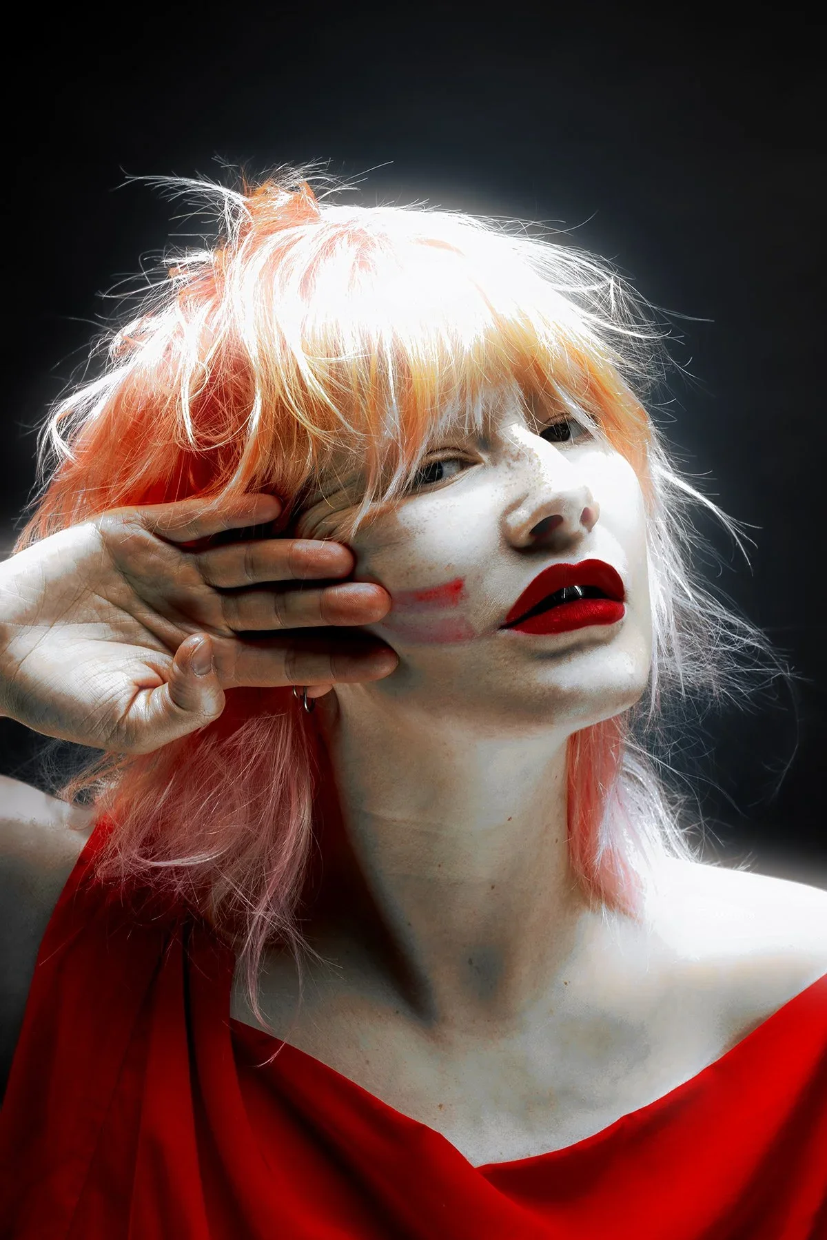 A person with pale skin and bright pink hair, wearing red lipstick, holding one hand to the side of their face, with a dark background.