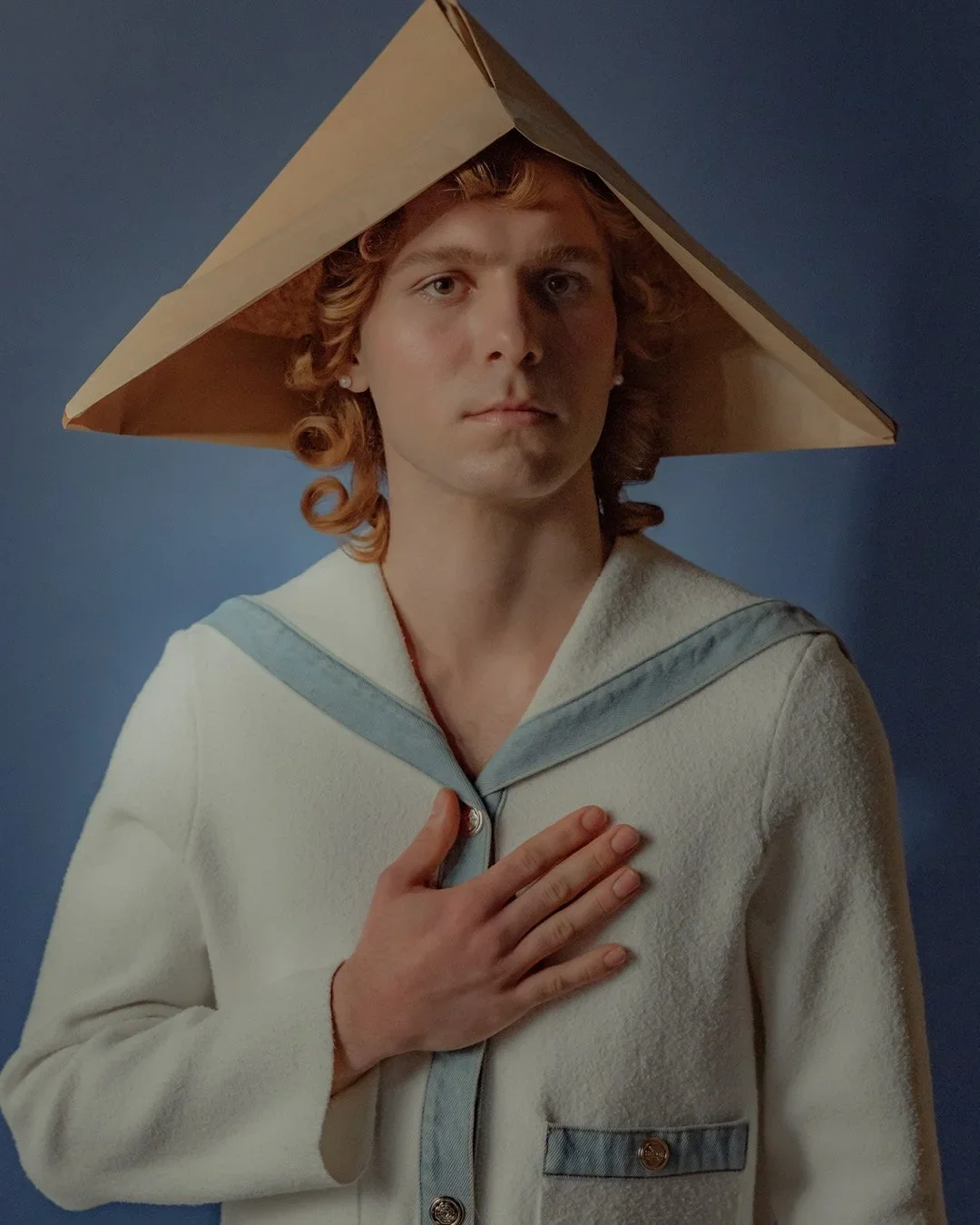 A person with curly red hair wearing a vintage sailor-style cream-colored coat with gray accents, a large triangular paper hat, and holding their hand over their chest against a blue background.