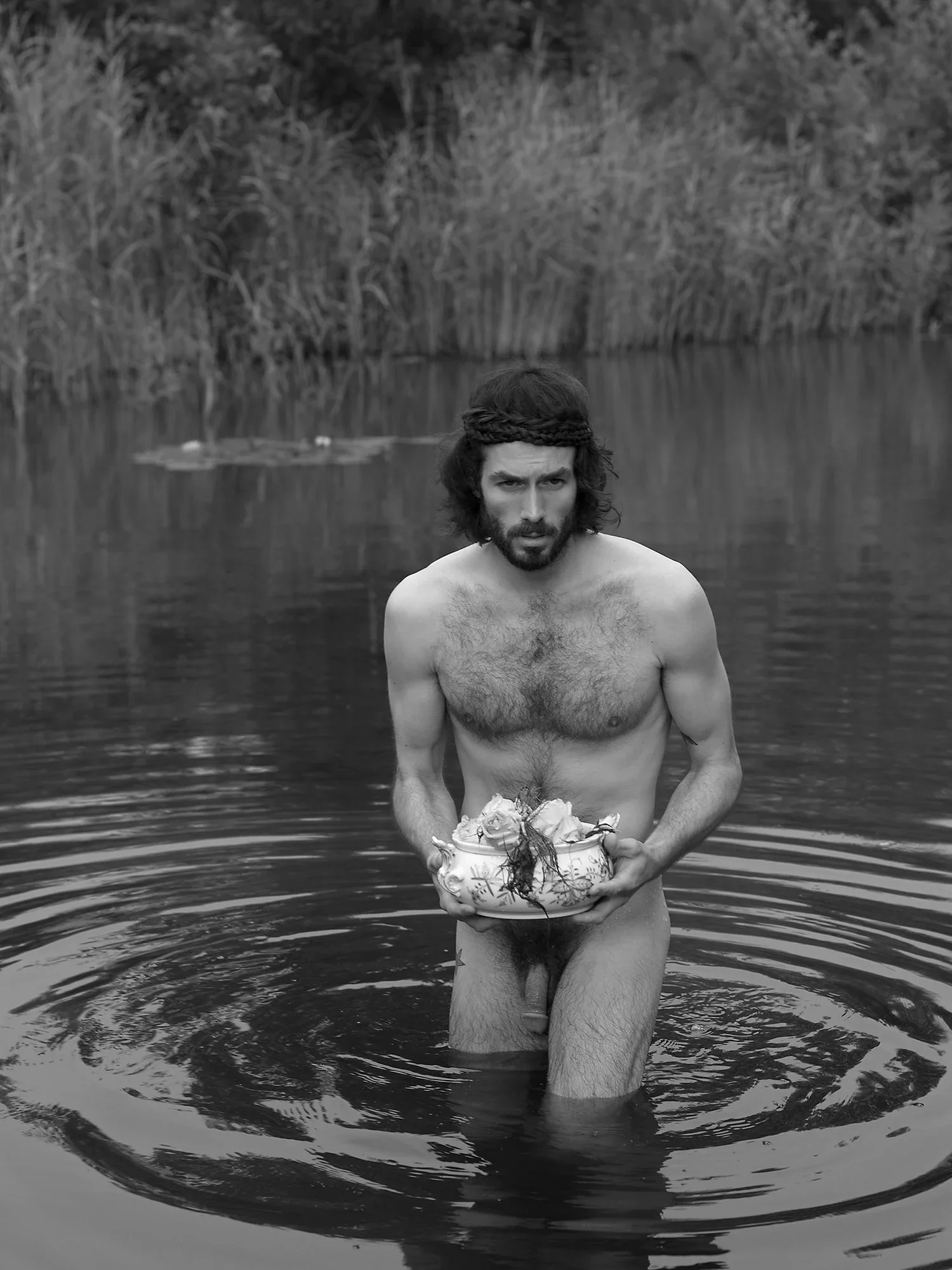 A shirtless man with long hair and a headband stands waist-deep in a body of water, holding a decorated bowl with flowers. The background features tall grass and reeds along the water's edge.