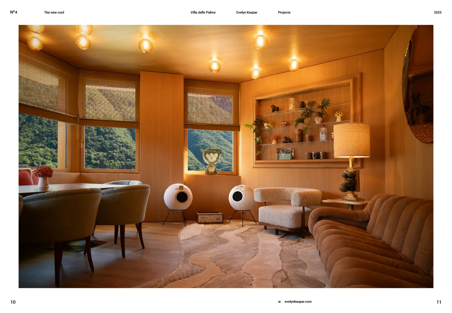 Living room with wooden walls, ceiling, and flooring, decorated with plants, shelves, and lighting. Large windows show green mountains outside. Furniture includes a beige couch, armchair, and dining table with chairs. Speakers and a lamp are also pre