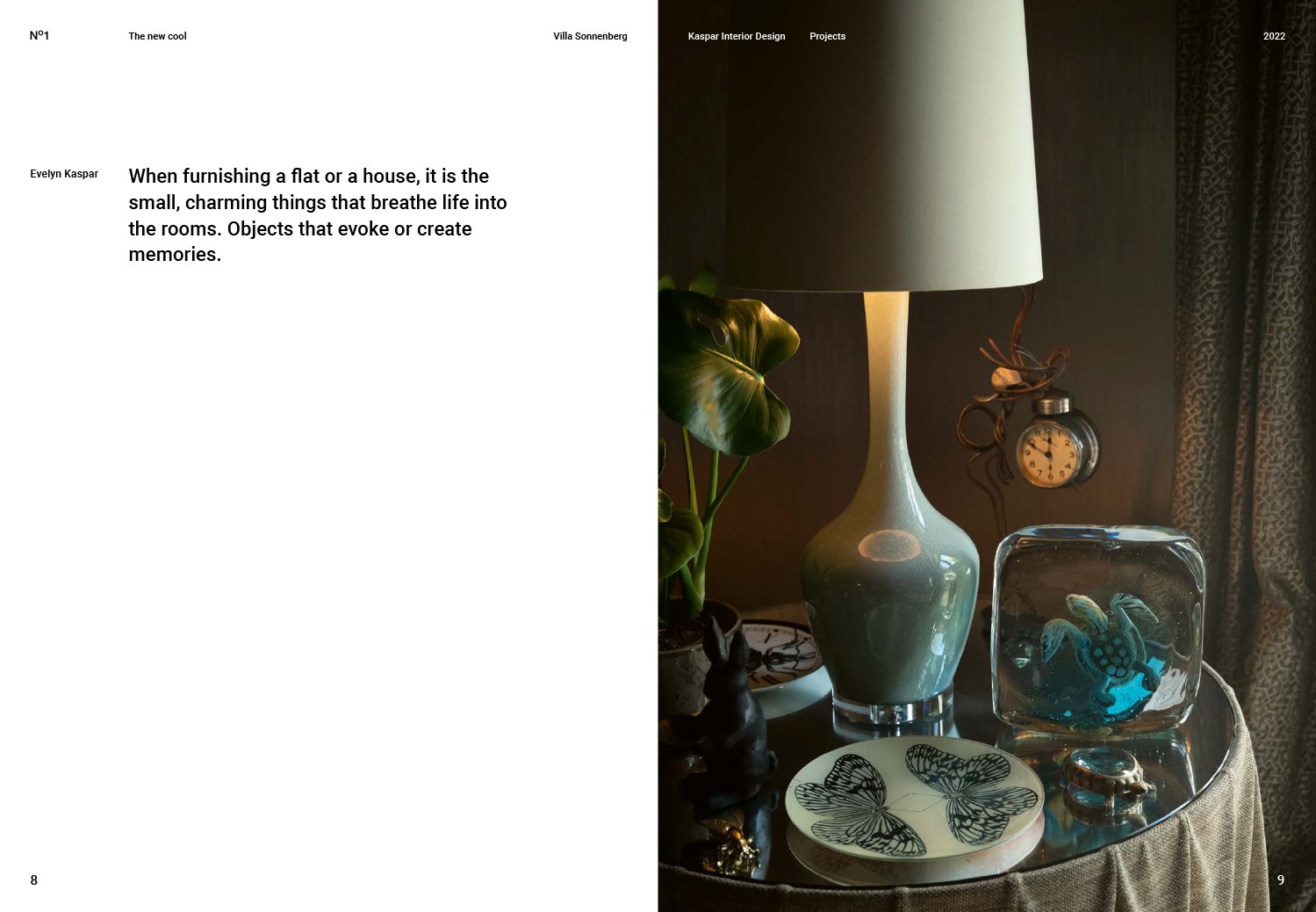 Close-up of a table lamp with a white shade on a side table, surrounded by decorative items including a glass turtle figurine, a black rabbit figurine, a plate with butterflies, a small clock, and a plant with large green leaves. Dark curtains are vi