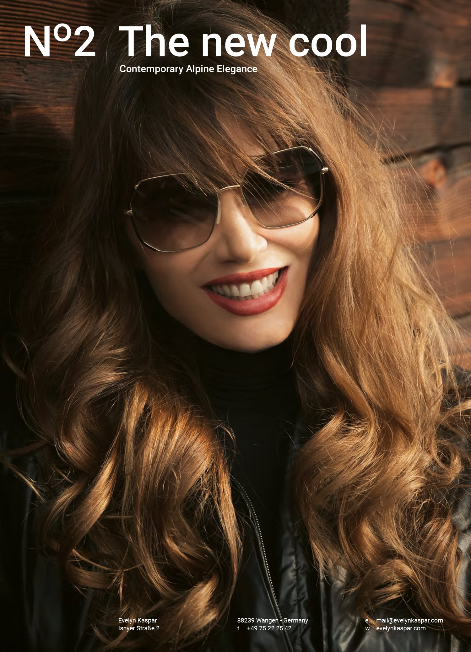 A woman with long, wavy red hair wearing sunglasses and a black leather jacket, smiling against a wooden background.