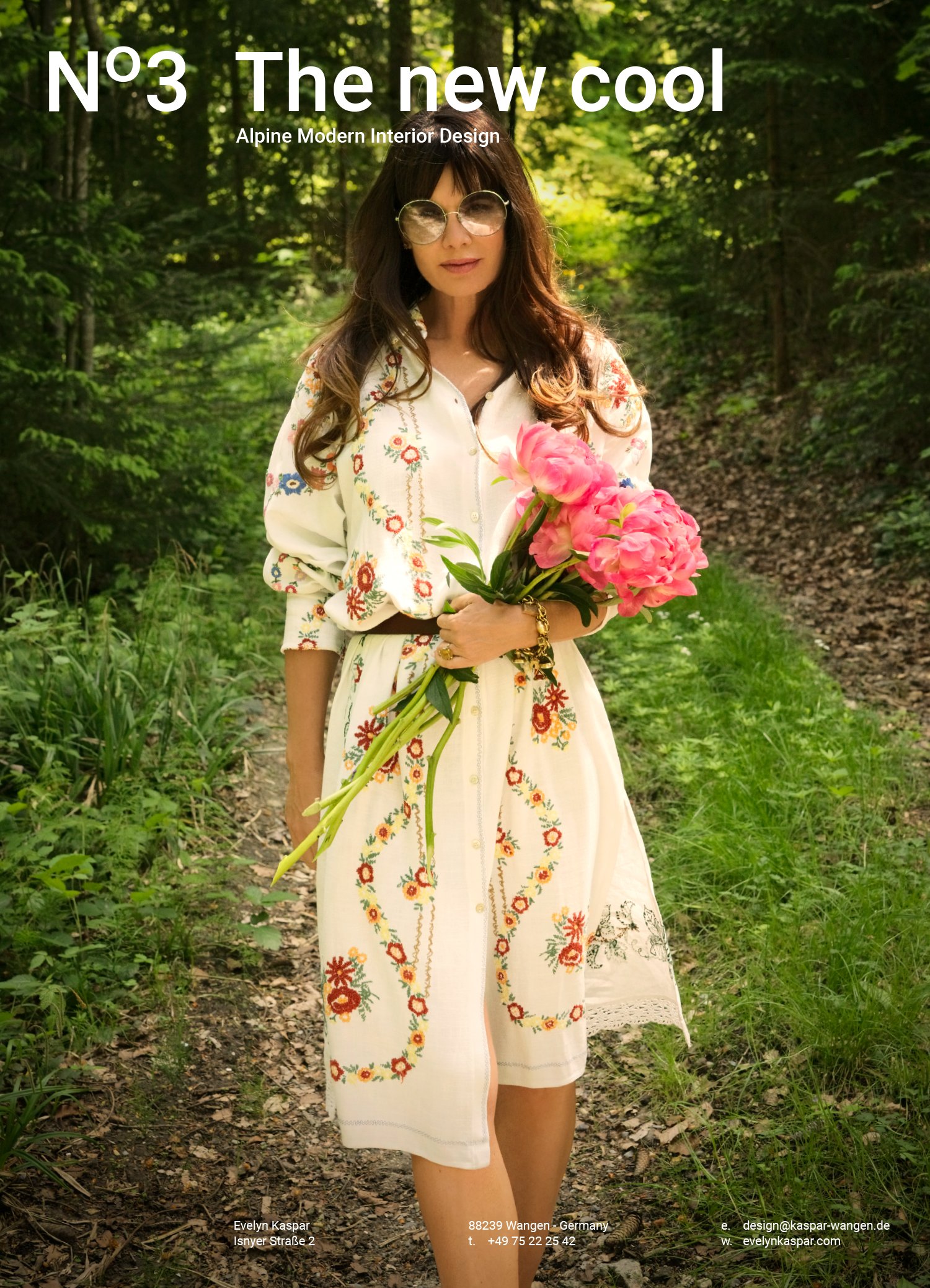 A woman wearing sunglasses and an embroidered white dress is standing on a forest path holding a bouquet of pink flowers.