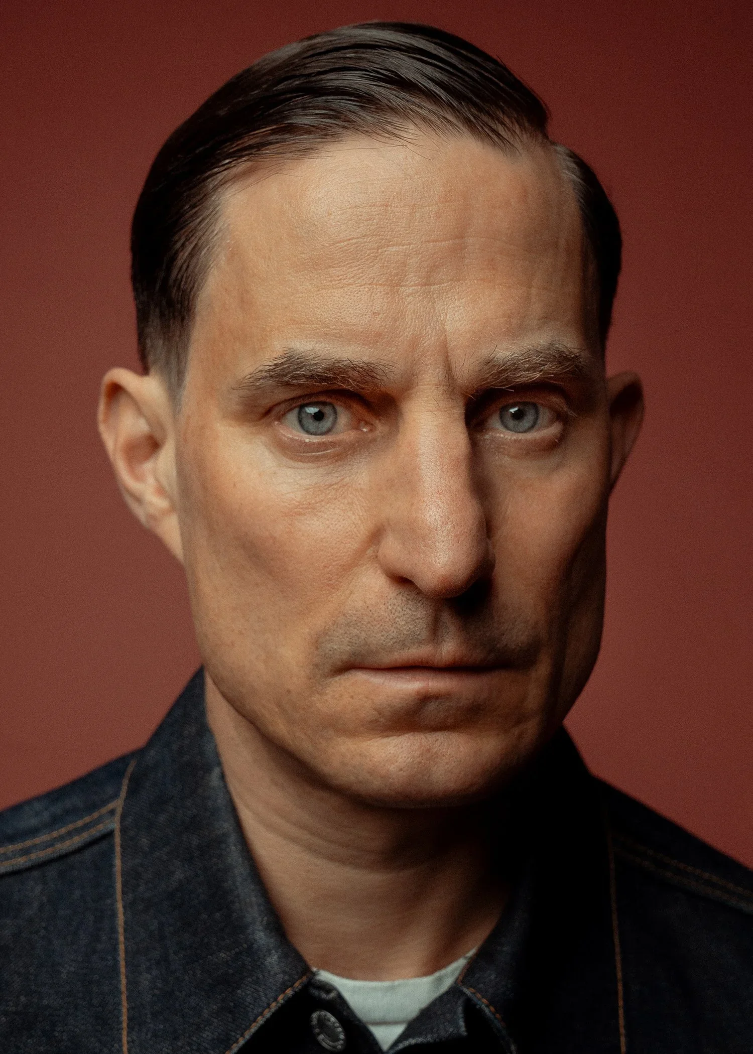 Close-up portrait of a man with blue eyes, slicked-back brown hair, wearing a dark denim shirt, against a reddish-brown background.