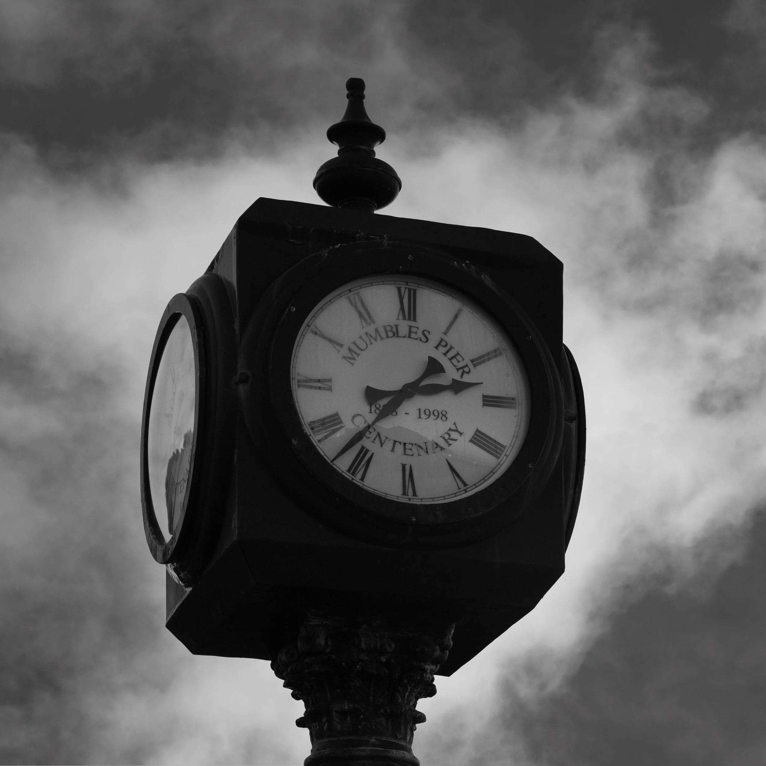 A black and white photo of a vintage clock tower with a cloudy sky in the background. The clock face reads 2:13 and is inscribed with 'Mumbles Pier Centenary 1898 - 1998'.