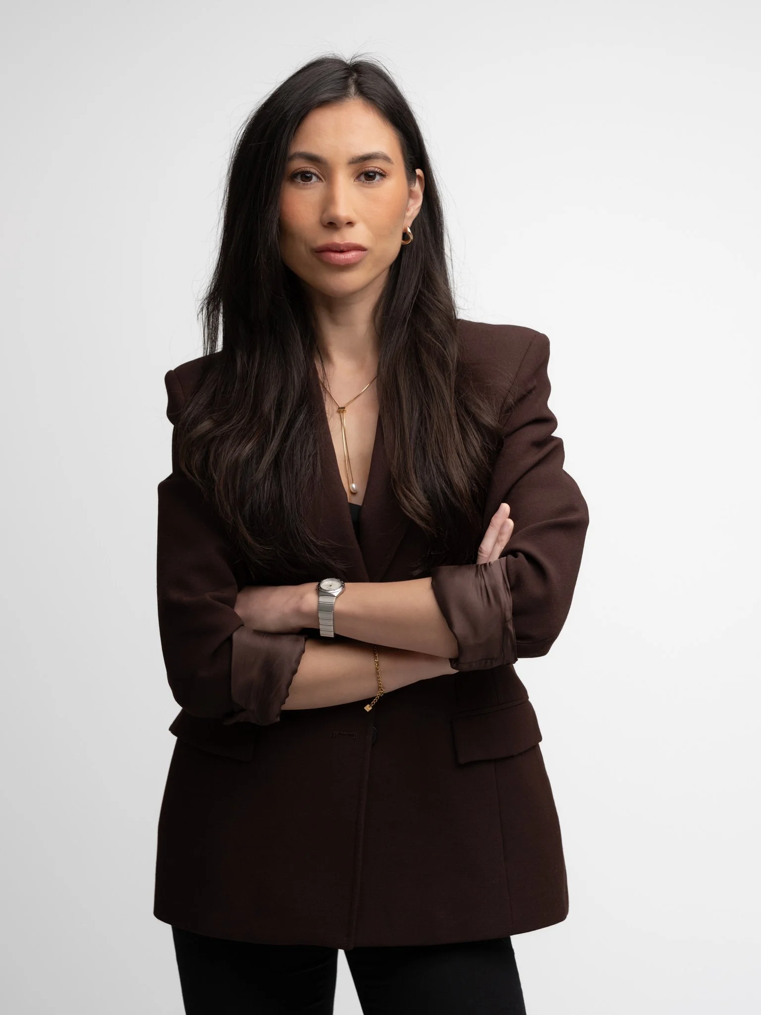 A woman with long dark hair, wearing a dark brown blazer with rolled-up sleeves, a gold necklace, a watch, and earrings, standing with her arms crossed against a plain white background.