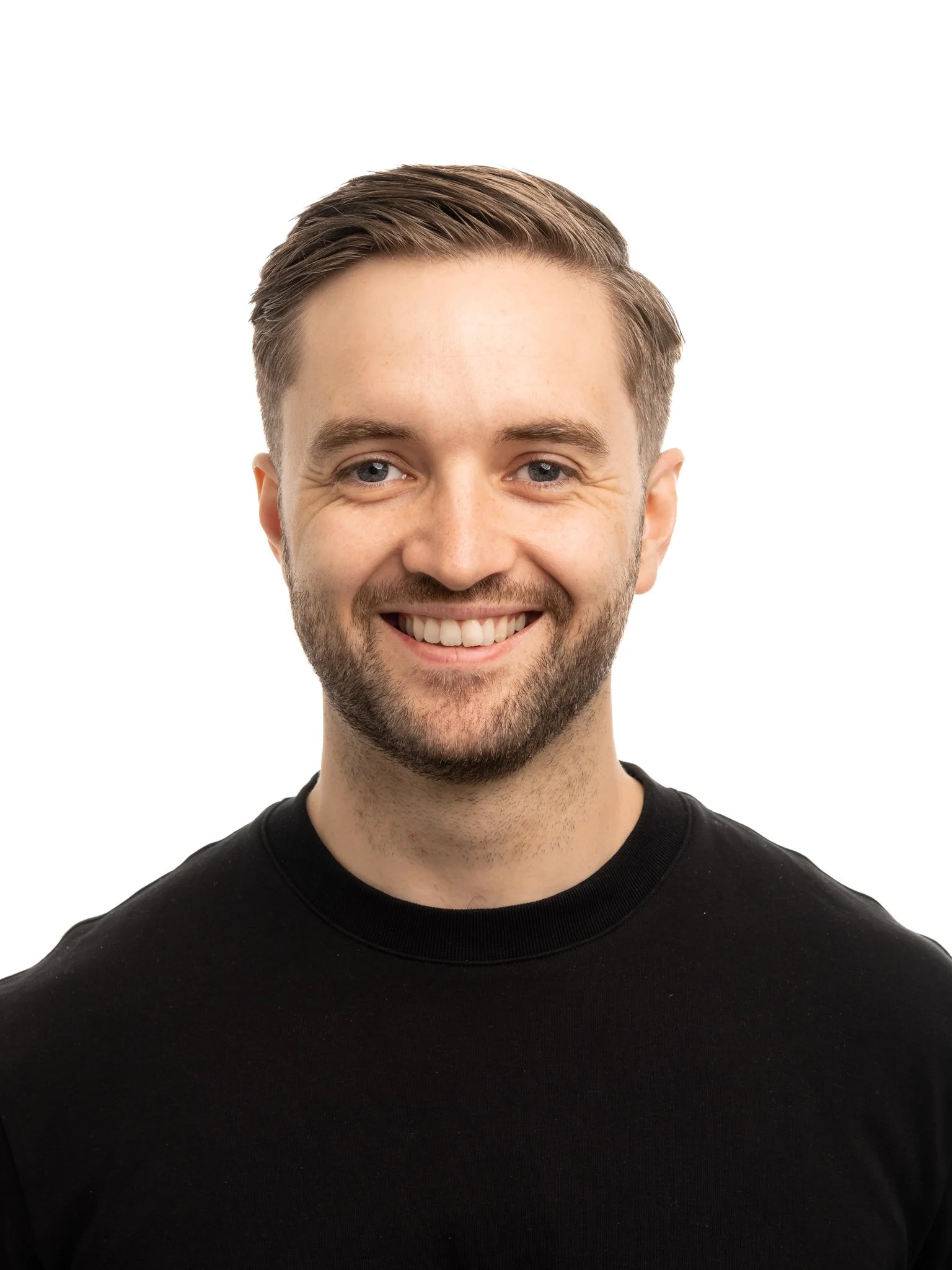A smiling young man with short brown hair and a beard wearing a black shirt, posing against a white background.