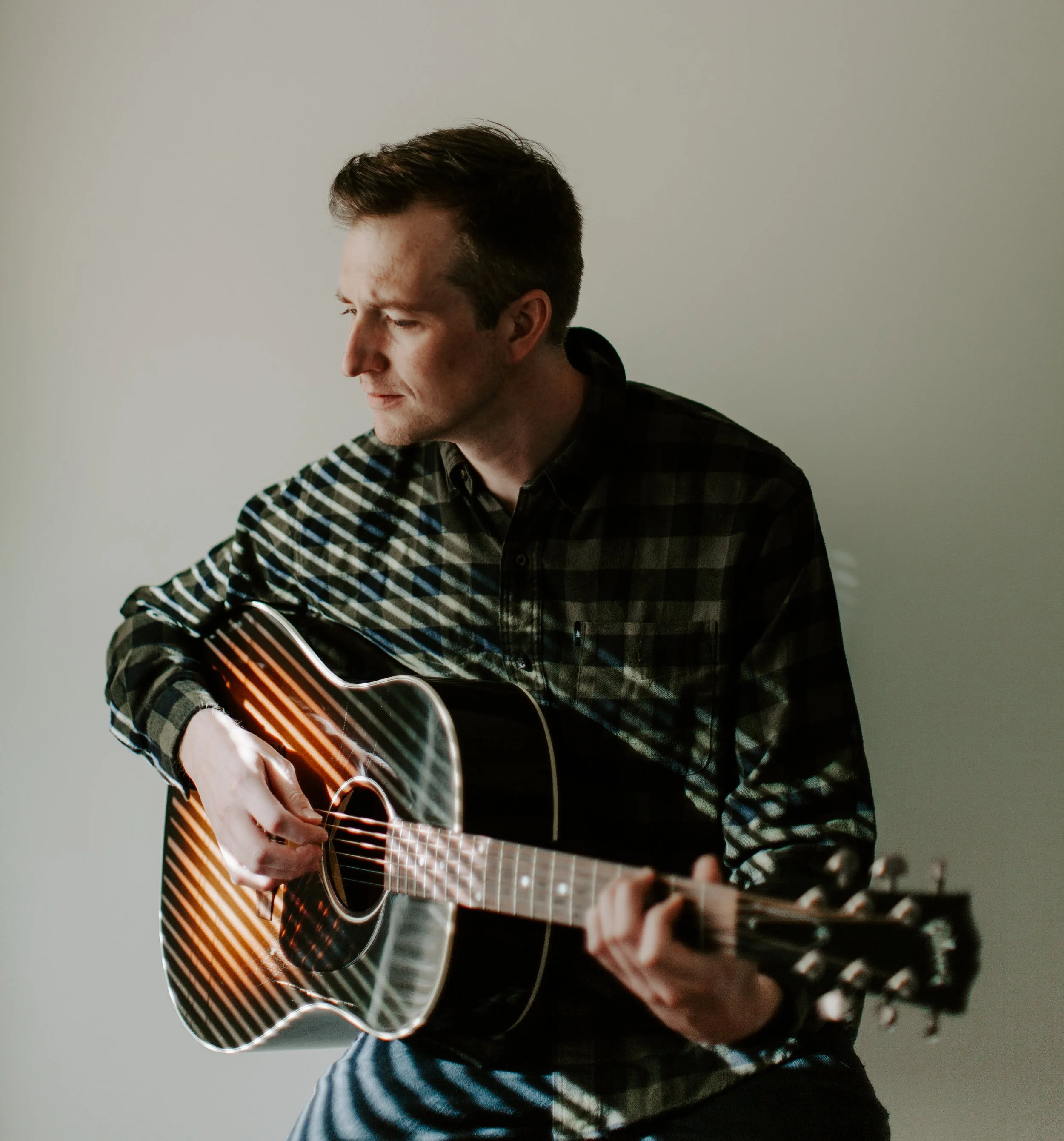 A man wearing a black and green plaid shirt playing an acoustic guitar, sitting against a plain light-colored wall.