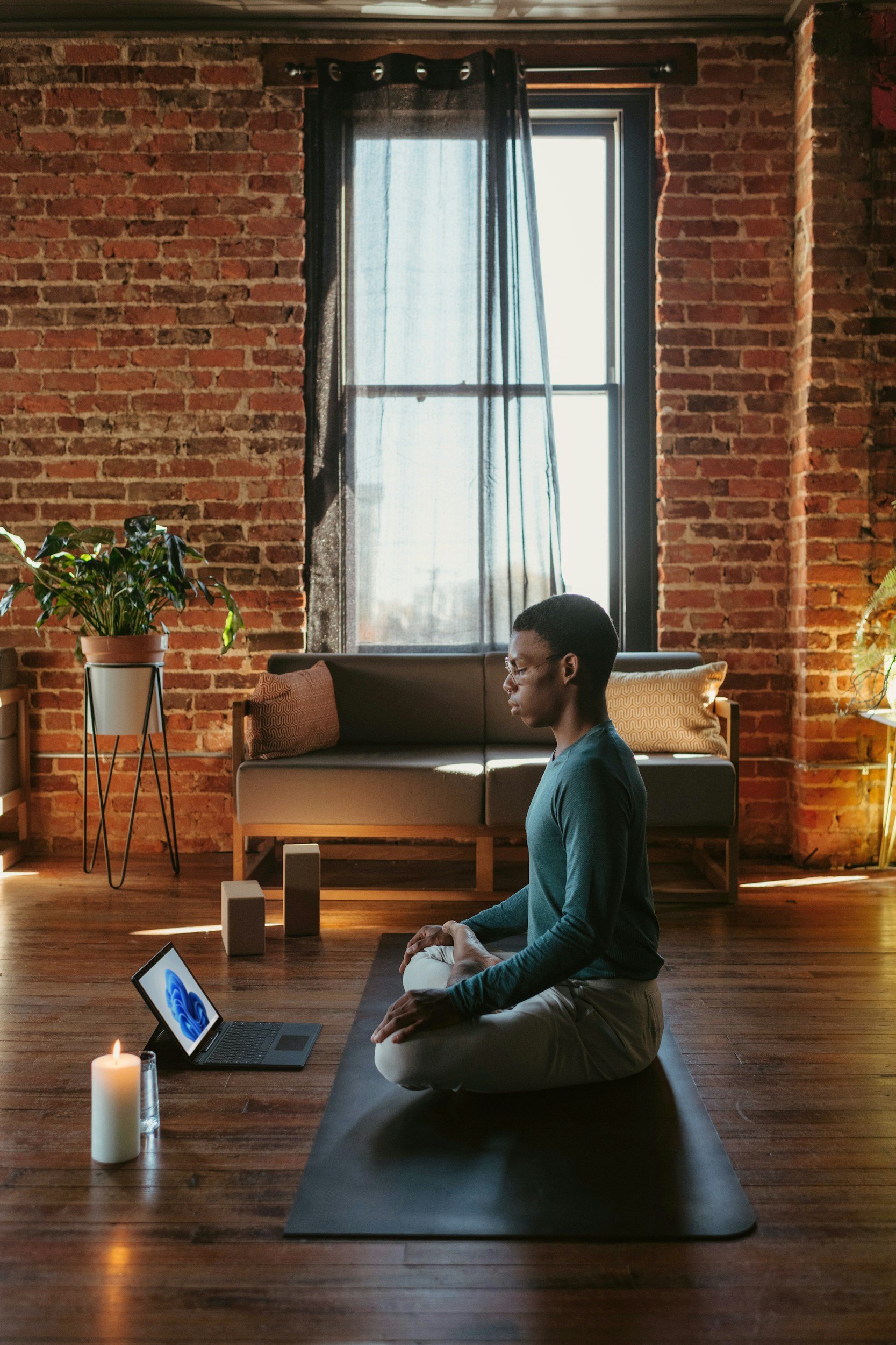 A person practicing yoga on a black mat in a cozy, sunlight-filled room with exposed brick walls, a window with sheer curtain, houseplants, and a laptop nearby.
