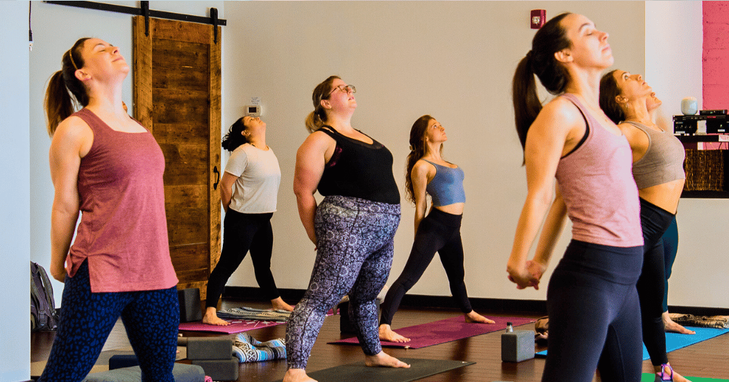 Group of women practicing yoga at Boston Yoga Union in Coolidge Corner, standing with hands behind back in a pose.