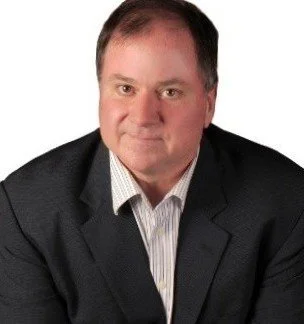 A man in a dark suit jacket and light dress shirt posing against a plain white background.