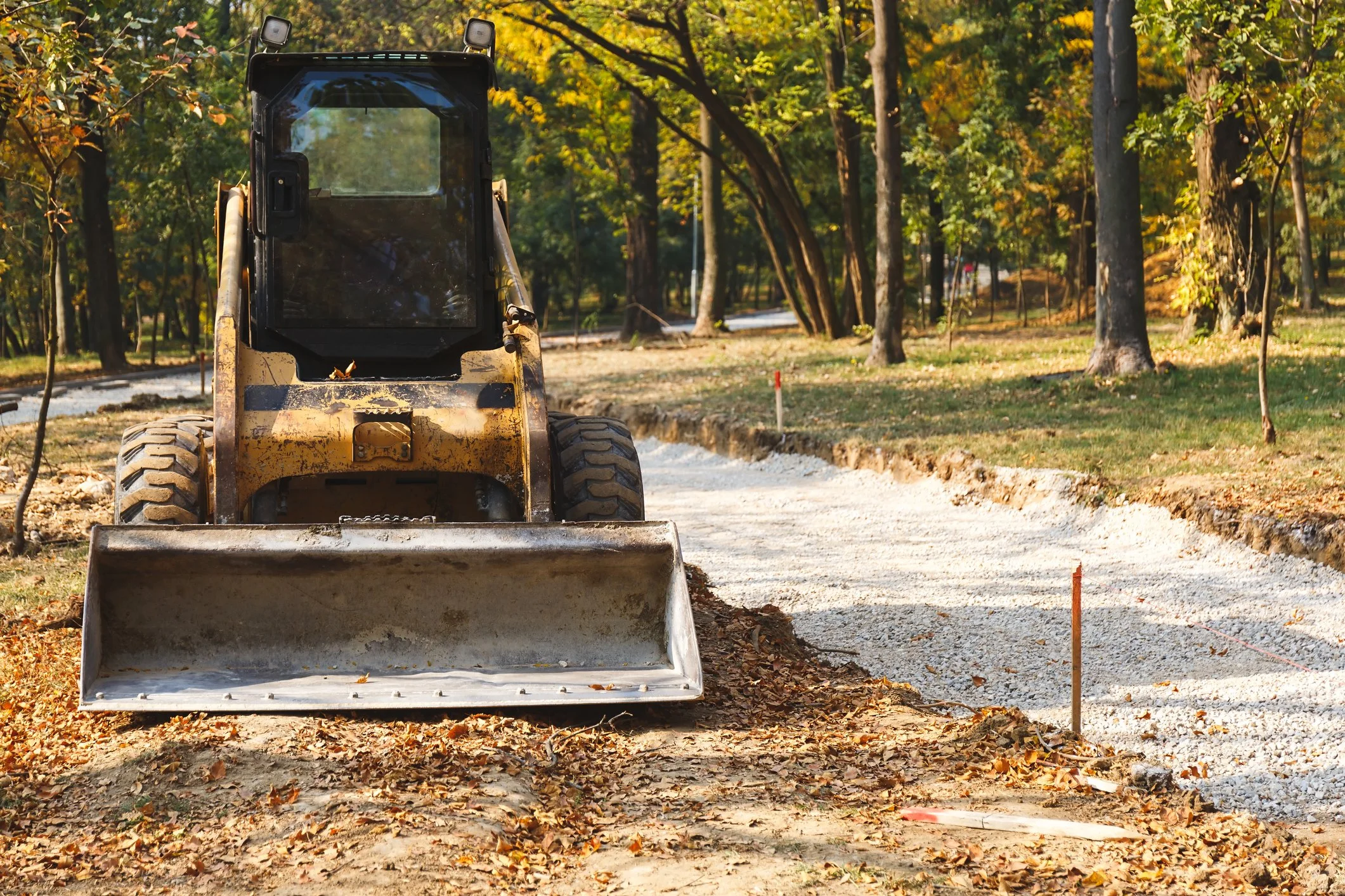 A construction vehicle working on a gravel pathway in a park surrounded by trees with fall foliage.