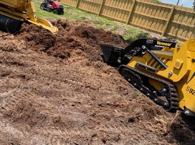Construction site with a small yellow bulldozer moving dirt, fenced backyard, and another tool in the background.