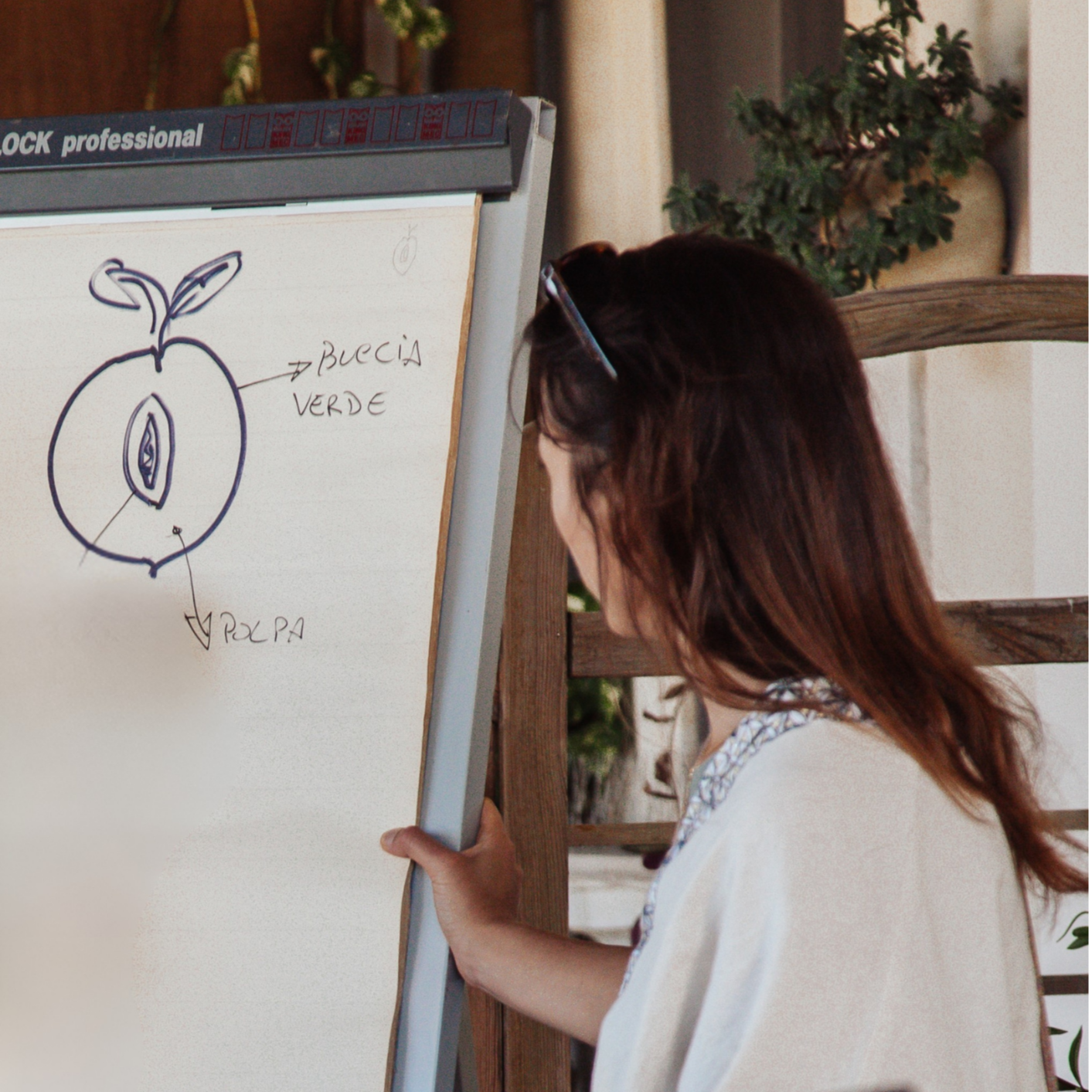 A woman standing outdoors, looking at a whiteboard with a hand-drawn diagram of a peach, labeled in Italian. The diagram includes the peach's parts marked as 'buccia verde' (green peel) and 'polpa' (flesh).