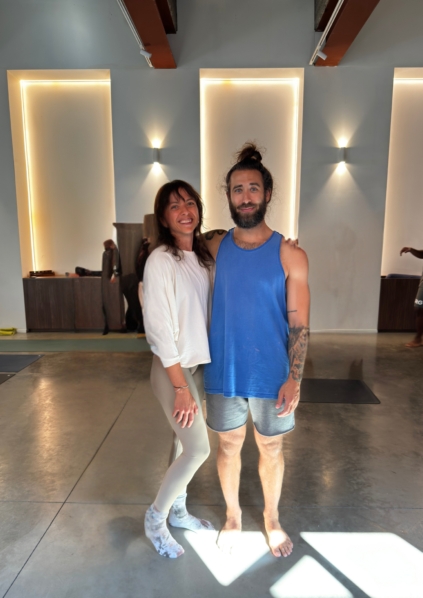 A smiling woman and man standing together indoors. The woman is wearing a white top and light-colored pants, and the man is in a blue sleeveless shirt and gray shorts. The setting appears modern with wall-mounted lights and a polished concrete floor.