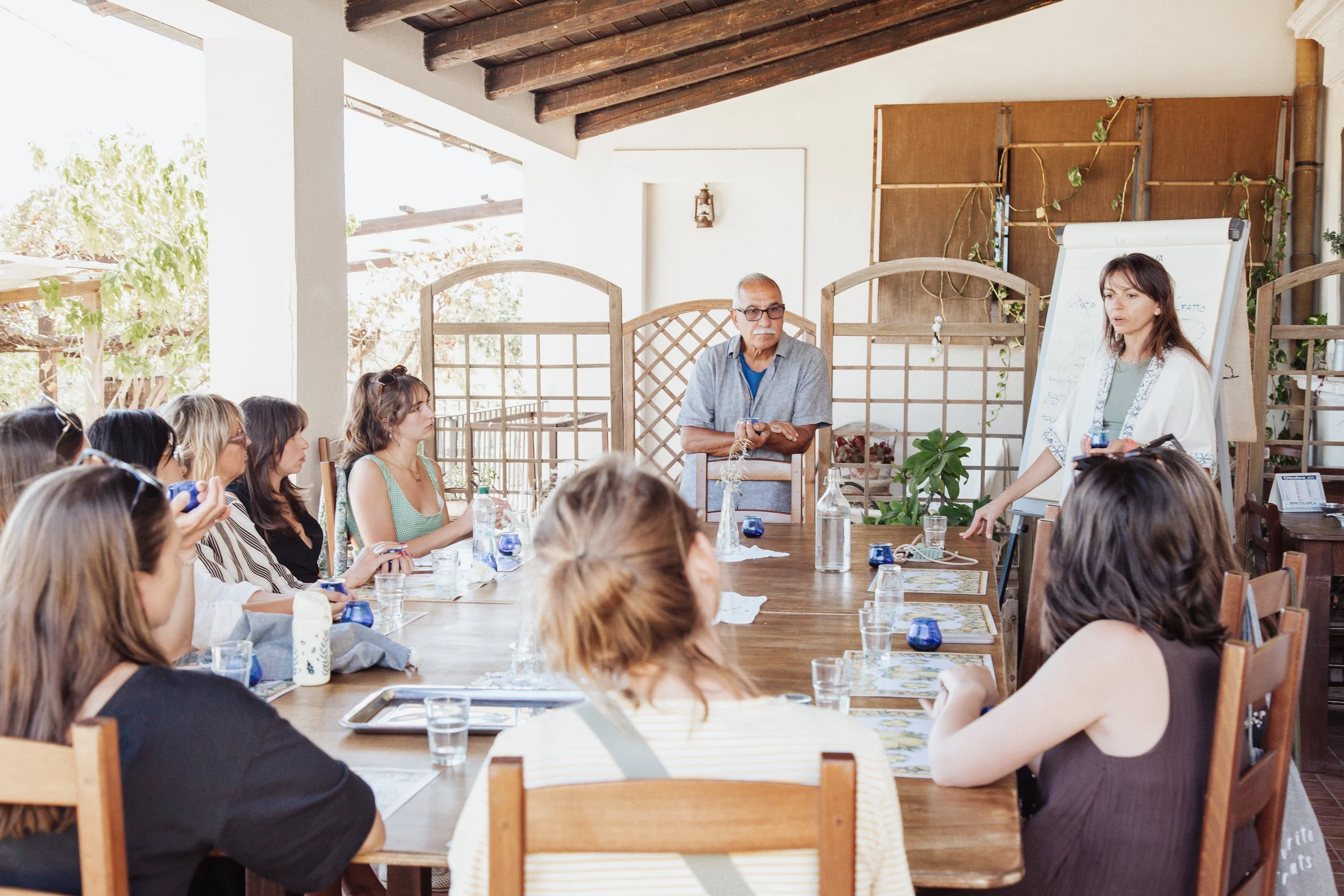 A woman standing by a whiteboard giving a presentation to a group of people sitting around a large wooden table in a bright, airy room.