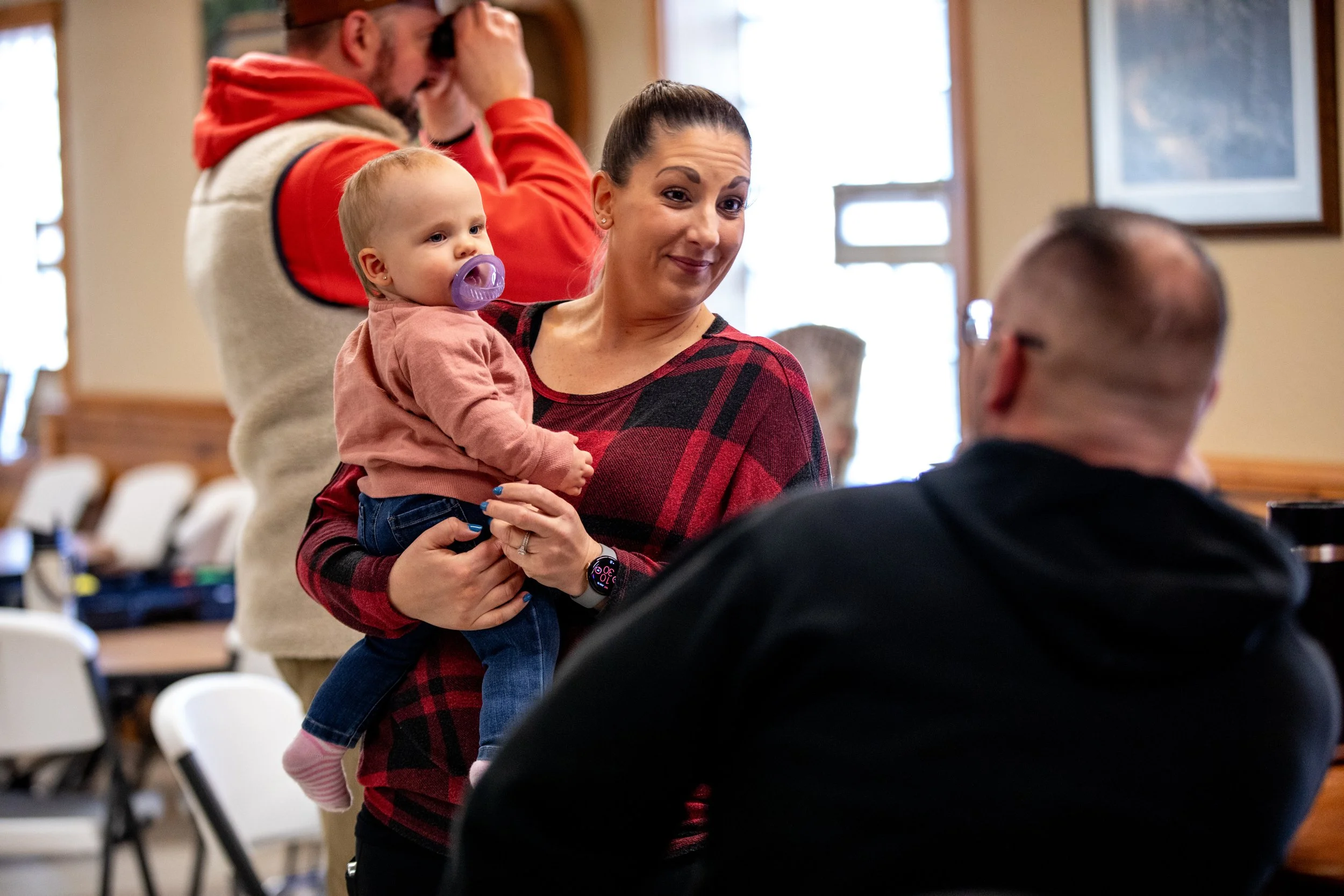 A woman holding a baby with a purple pacifier, engaging in conversation with a man in a black hoodie inside a room with wooden walls and framed pictures. A man in a red hoodie and a woman in a beige vest are in the background.