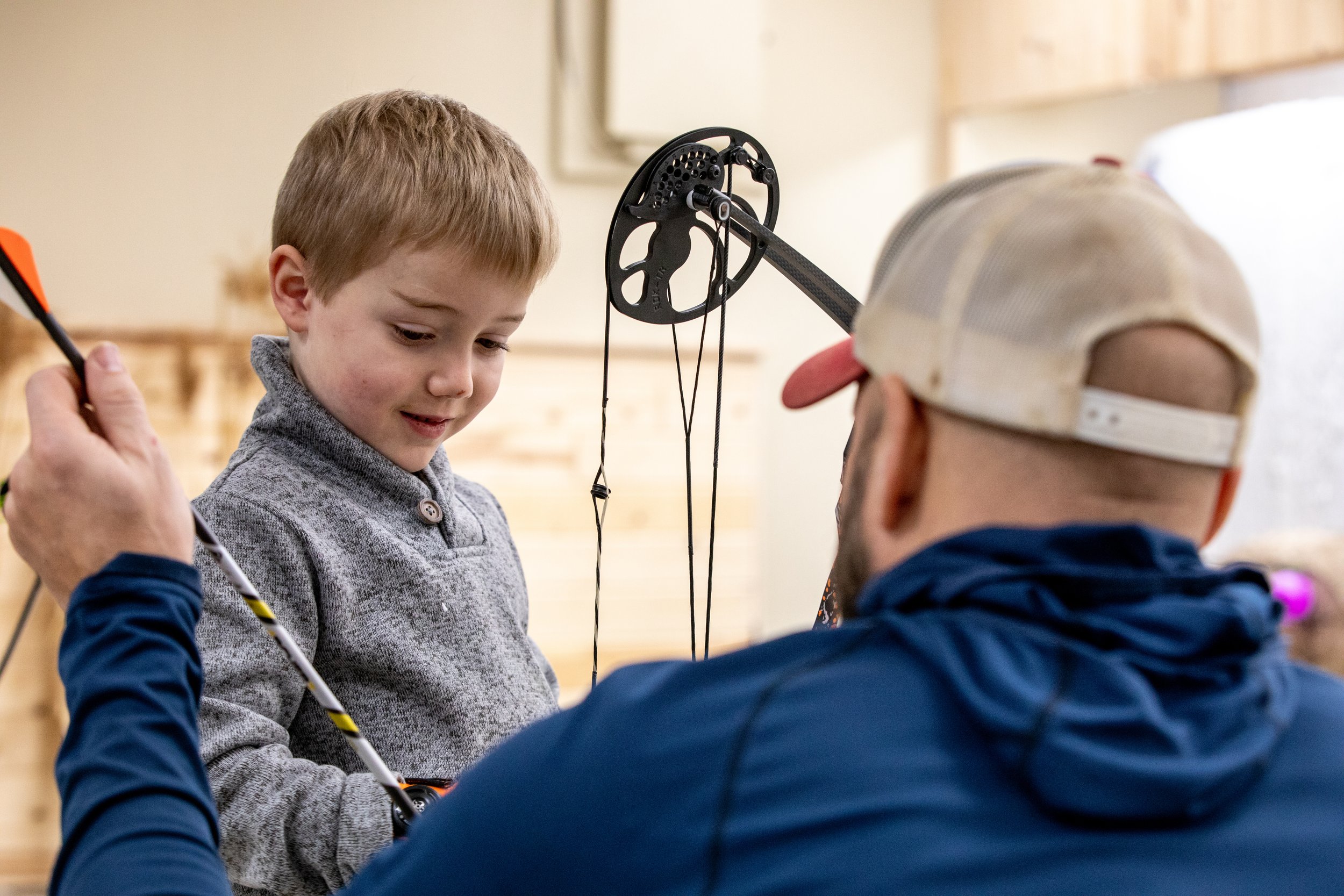 A young boy receiving archery instructions from a man in a cap in an indoor archery range.