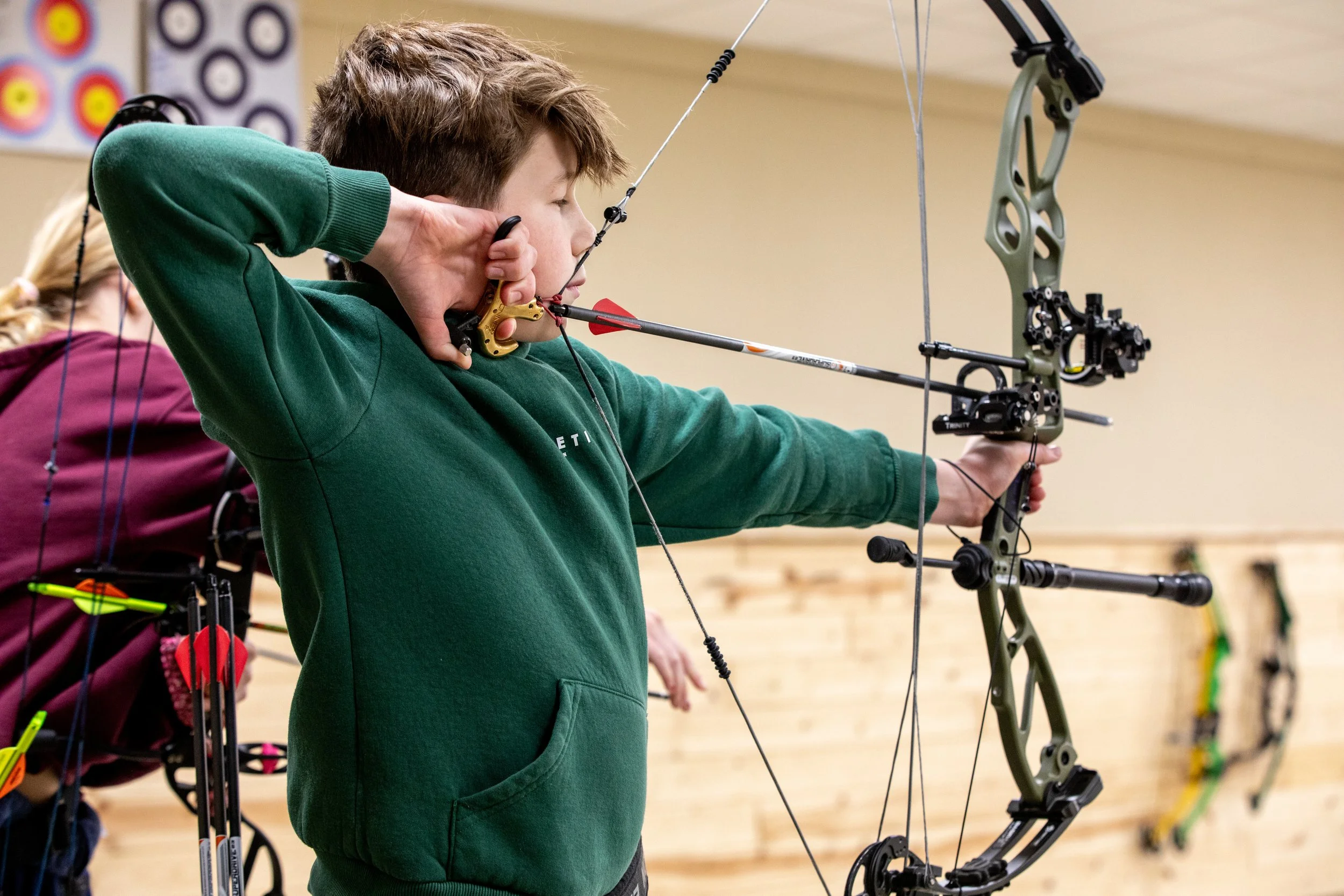 A young boy practicing archery indoors, aiming with a bow and arrow.