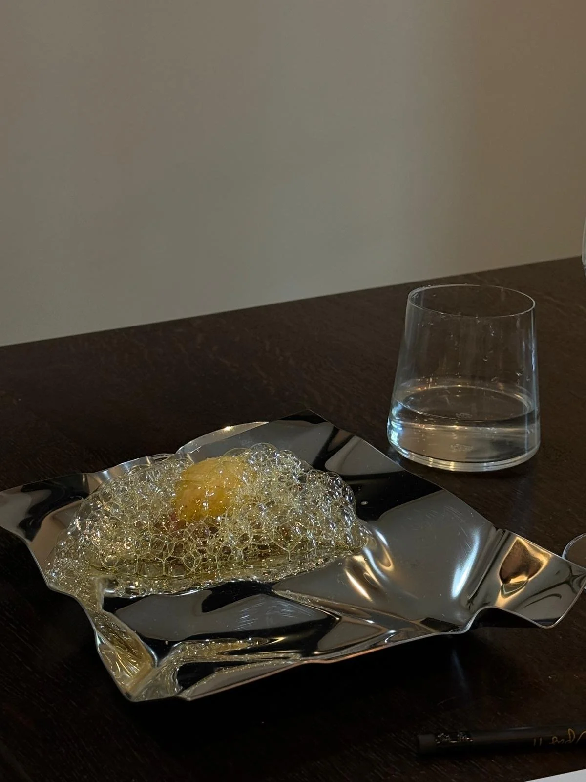 A piece of food covered in bubbles on a shiny, crinkled metallic plate, with a glass of water nearby on a dark wooden table.