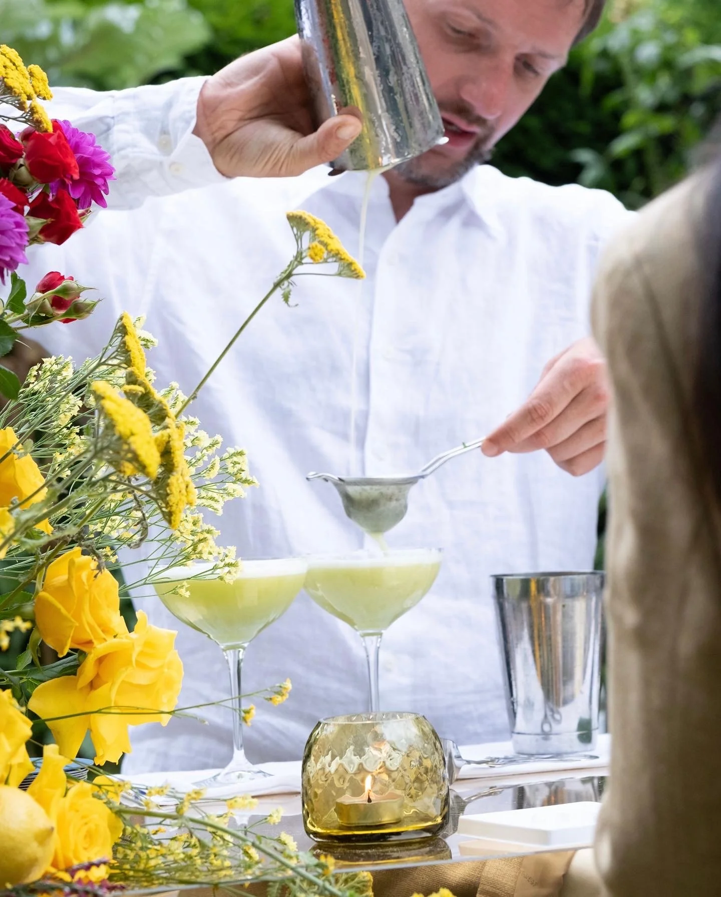 A man in a white shirt pours liquid from a metal shaker into a strainer over two coupe glasses filled with a green cocktail, with colorful flowers and candles on the table.
