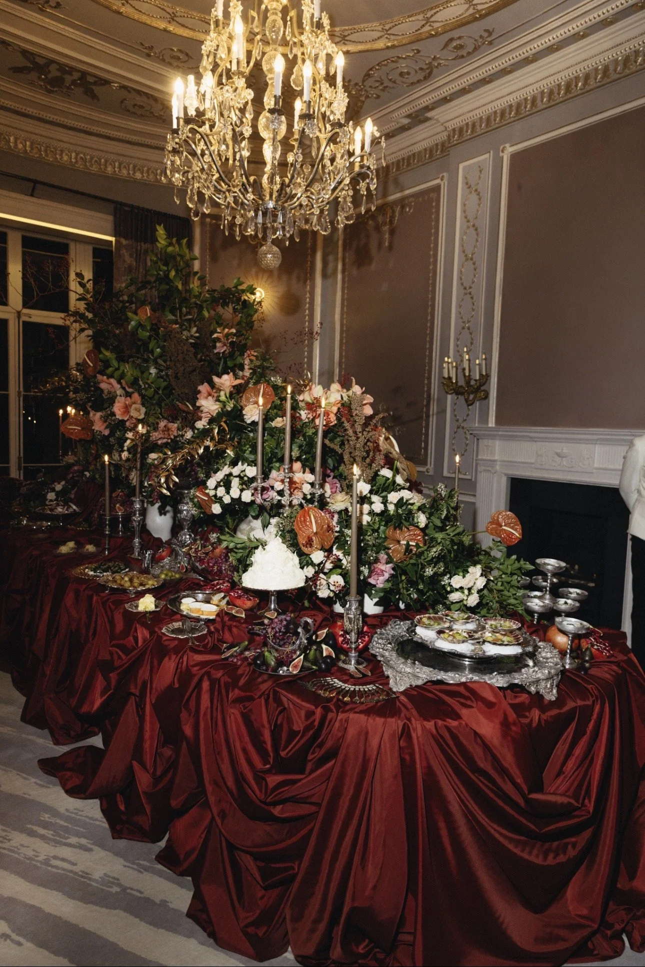 An opulent feasting table featuring dark red table draping, extravagant silverware holding oysters, scallops, salmon en croute, also featuring candles and flowers also including a live Jamon Iberico carving station