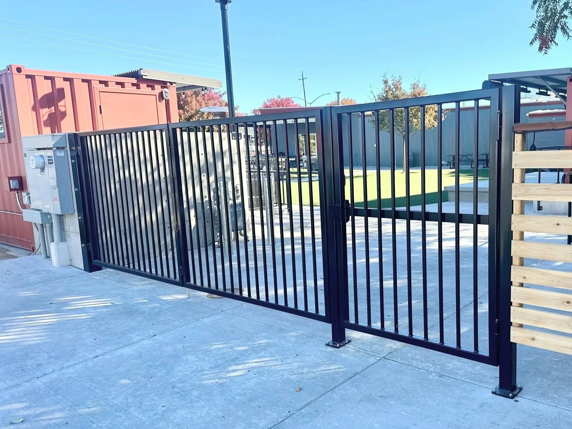 A black metal fence with a gate in a concrete-paved outdoor area, with a pink storage container, utility equipment, and trees with fall foliage in the background.