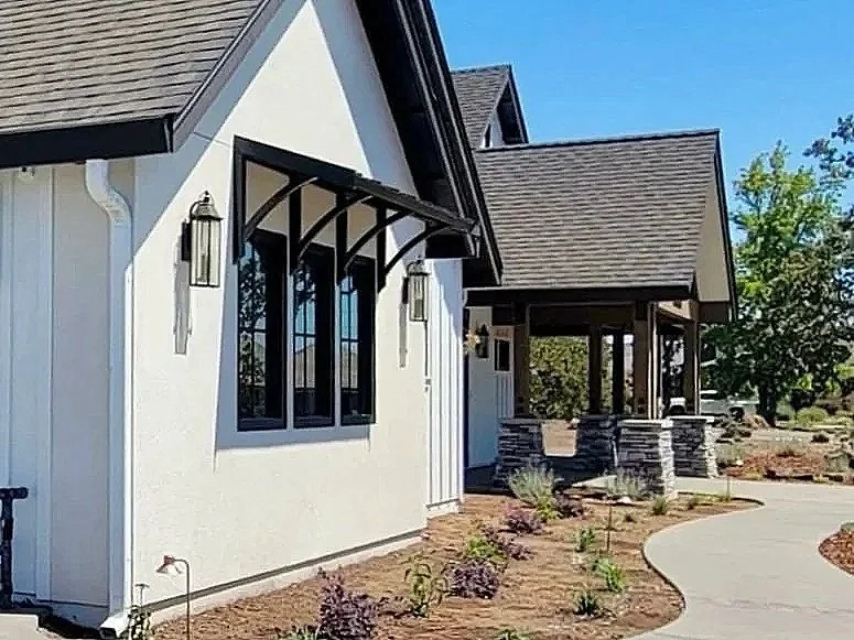 Exterior of a modern house with white walls, black window frames, and a gabled roof. The front yard has a curved concrete walkway and newly planted shrubs.