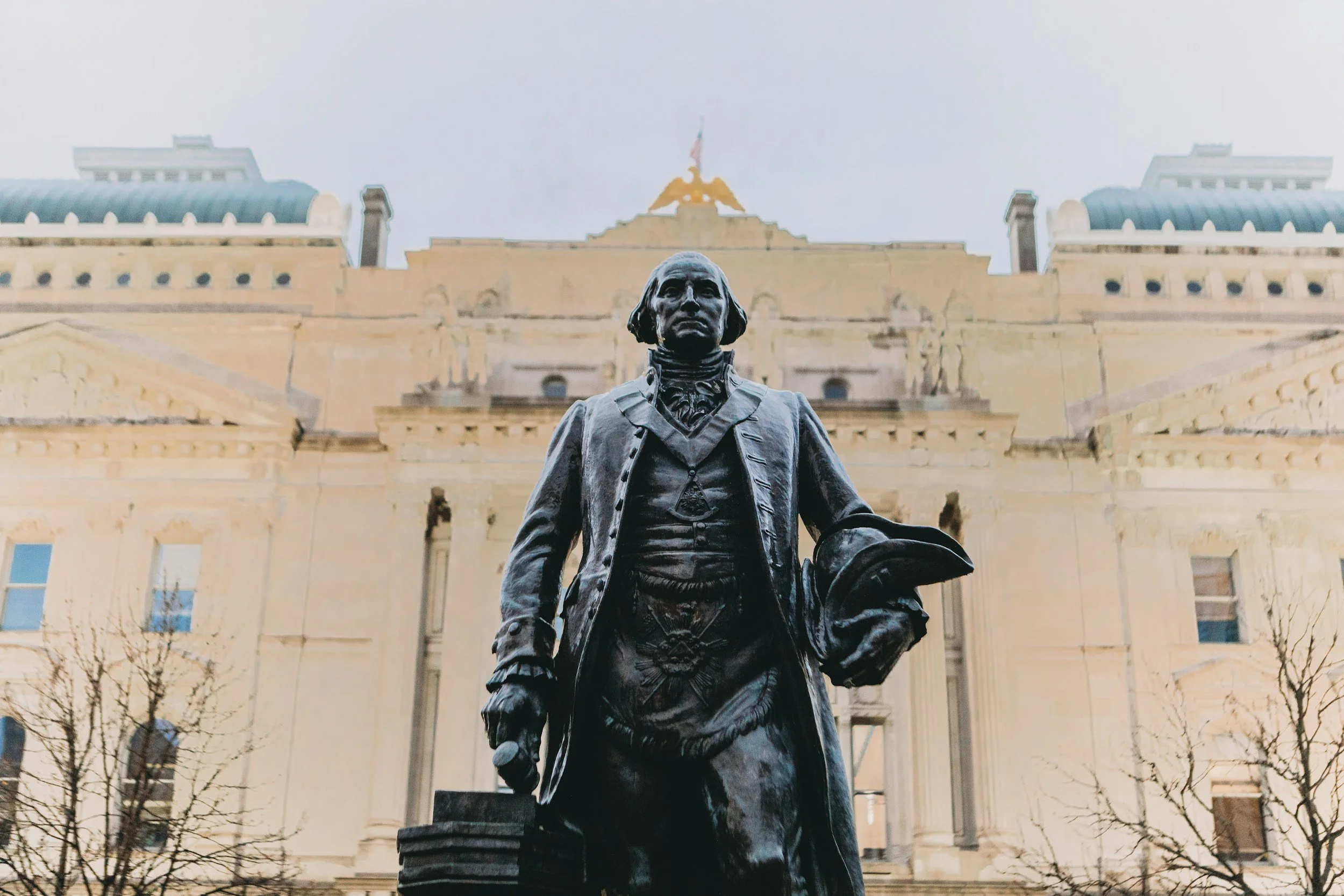 Statue of a man in 18th-century attire in front of a large government building with classical architecture, topped with a golden statue.