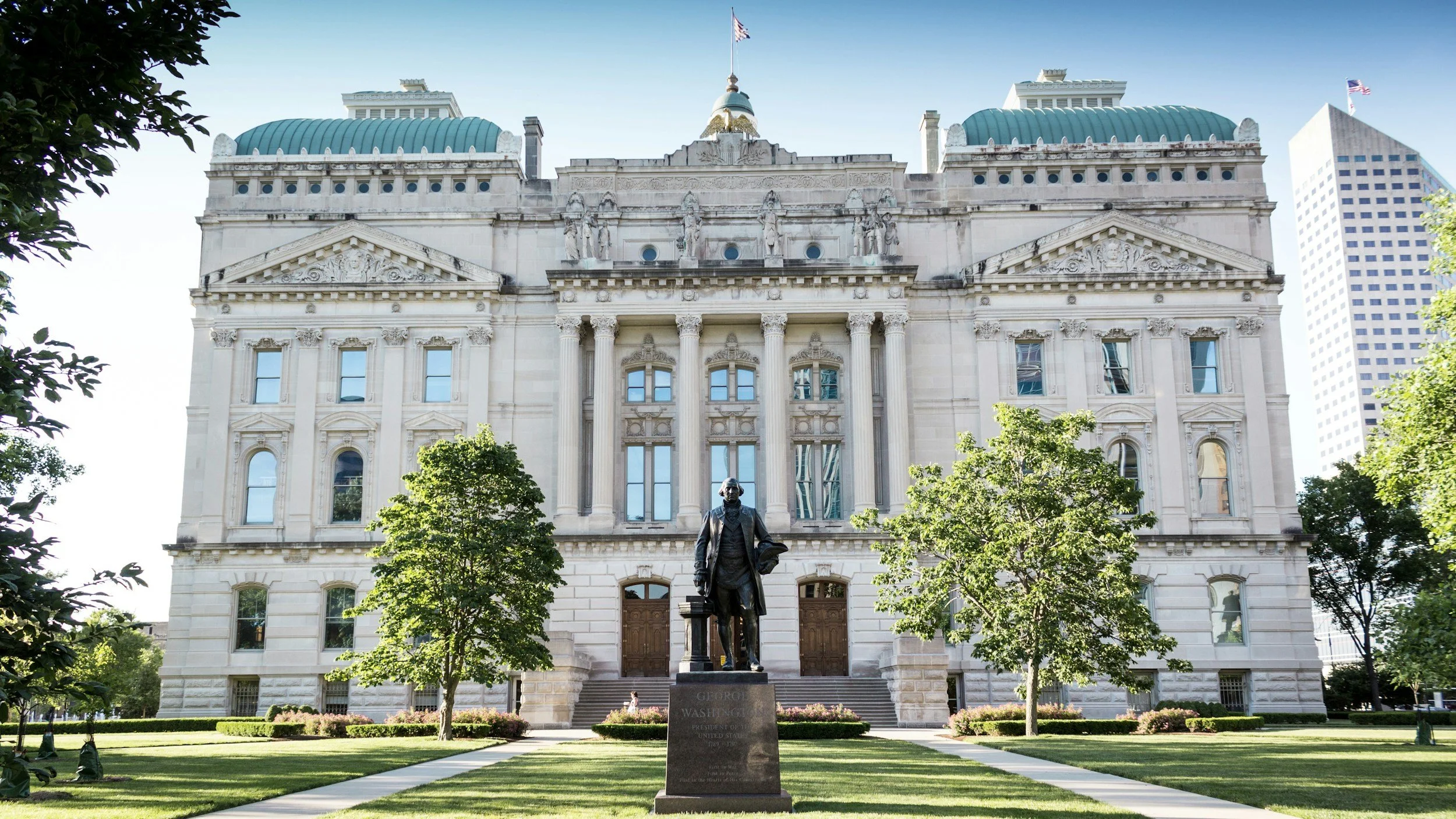 The Ohio Statehouse, a grand historical government building with classical architecture, white stone facade, columns, and statues, featuring a statue of George Washington in front, surrounded by trees and a well-kept lawn.