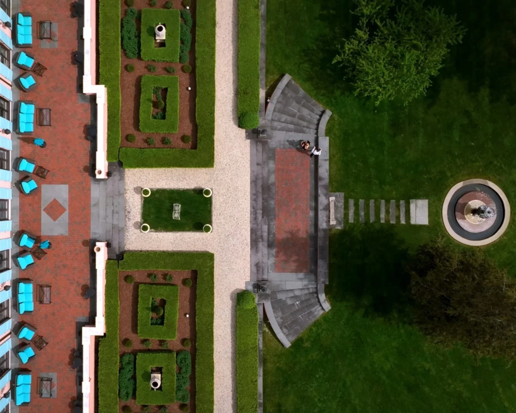 An aerial view of a landscaped outdoor space with a red-tiled rooftop area featuring blue seating, a fountain, green bushes, and walkways.