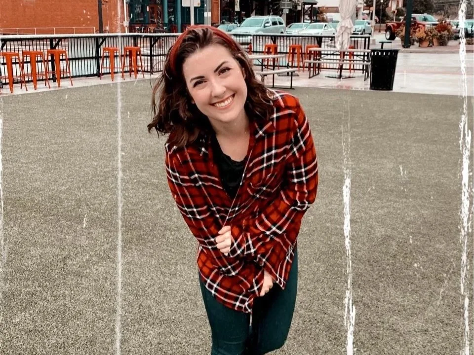 Forensic Science professor and author, Morgan Leslie, a young woman with curly brown hair, wearing a red plaid shirt, is smiling and leaning forward in an outdoor setting with orange chairs and tables in the background.