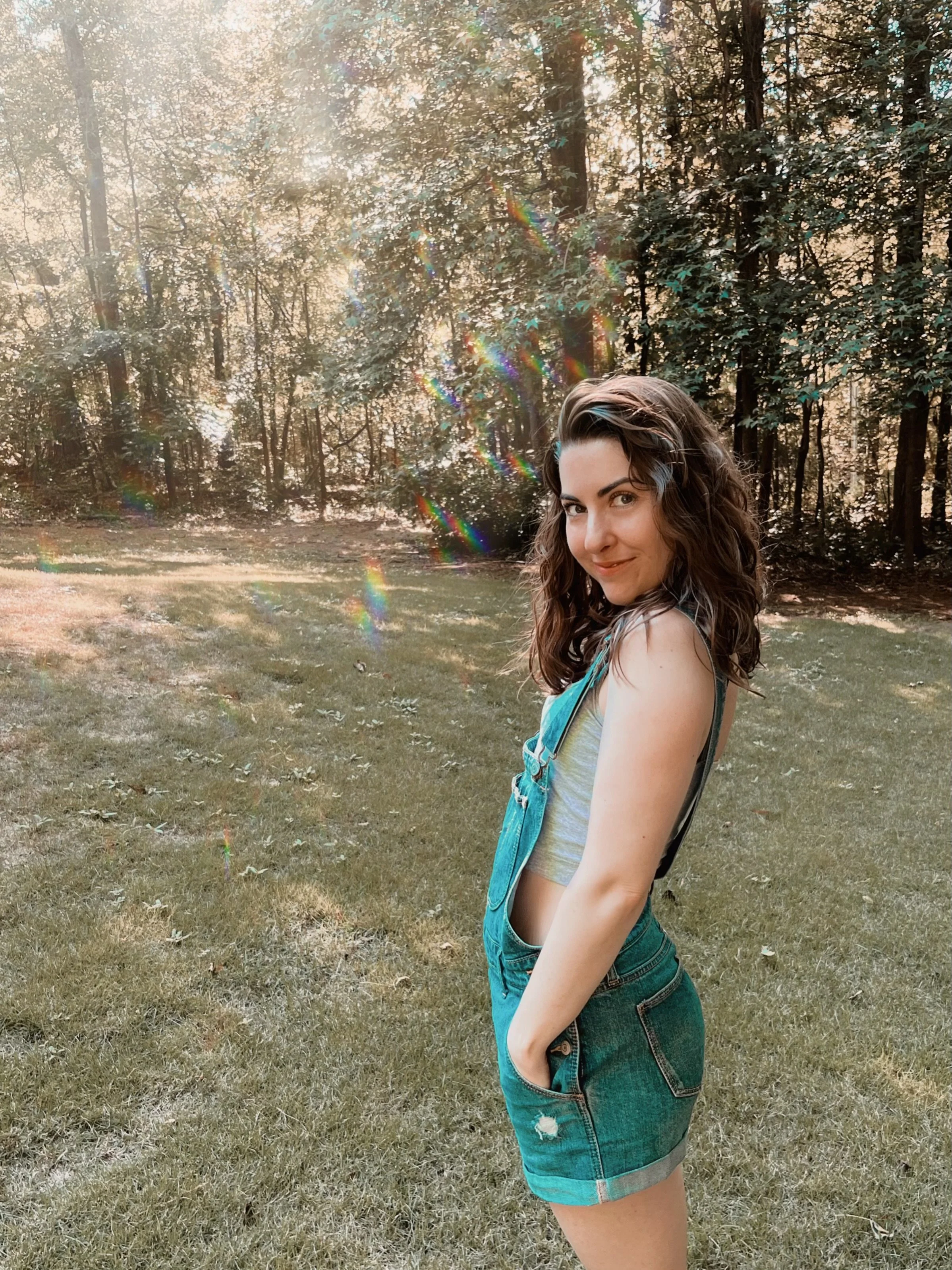 Morgan Leslie, a young woman with brown curly hair and a smile, standing outdoors in a grassy field with a forest of trees in the background, sunlight filtering through the leaves, and rainbow lens flare effects.