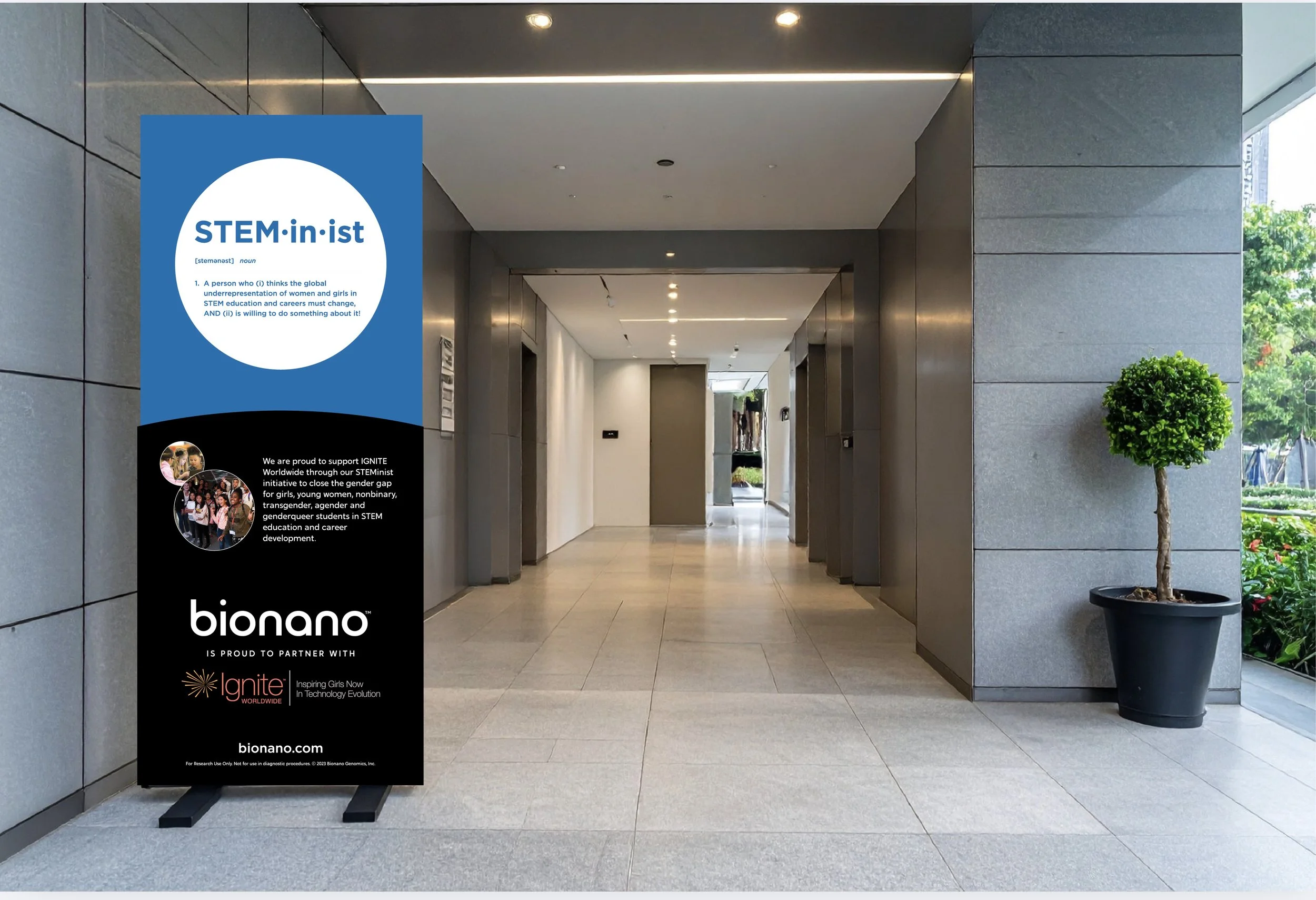 An indoor corridor features a sidewalk sign promoting STEM-in-ist and Bionano partnerships with Ignite Worldwide, with a potted tree on the right and elevators on the left, under ceiling lights, outside a glass wall with a view of greenery.