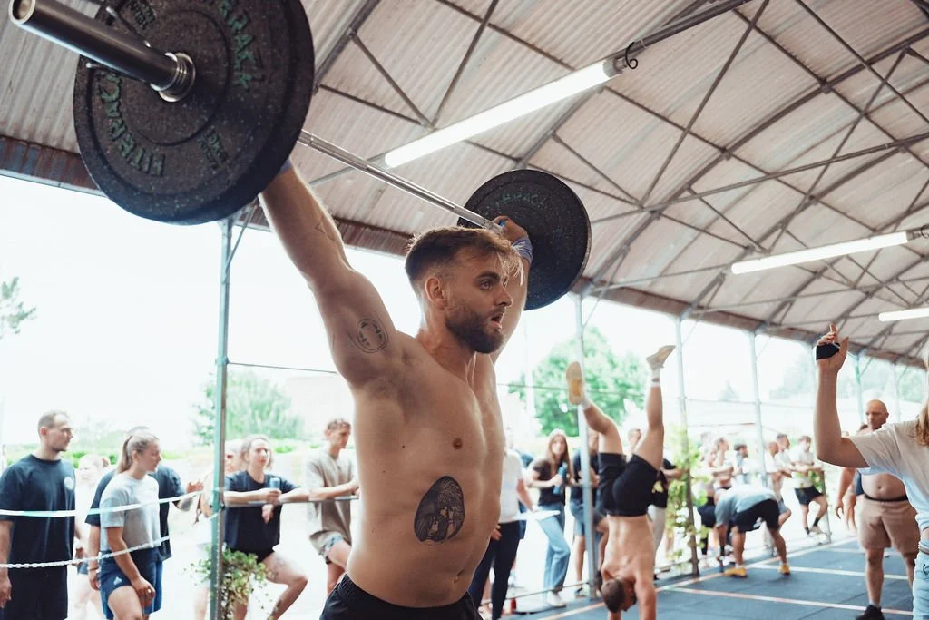 Un homme hommes dans une salle de sport, soulevant une barre avec des haltères au-dessus de sa tête, avec plusieurs autres personnes en train de faire des exercices en arrière-plan.