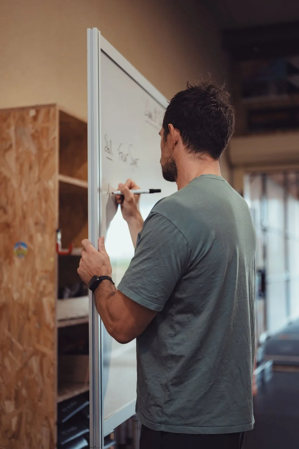 Homme en vêtements décontractés écrivant sur un tableau blanc dans un espace intérieur avec des étagères en bois derrière lui.