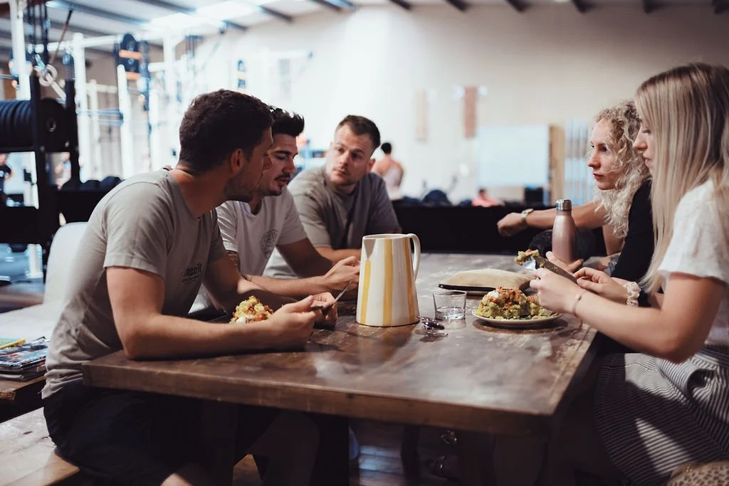 Groupe de jeunes adultes assis à une table en bois, partageant un repas dans un café ou un restaurant, avec des plats et une bouteille d'eau.