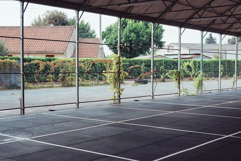 Espace vide de sport ou d'activité en intérieur avec un sol en caoutchou, clôture métallique et vue sur des maisons et arbres en arrière-plan.