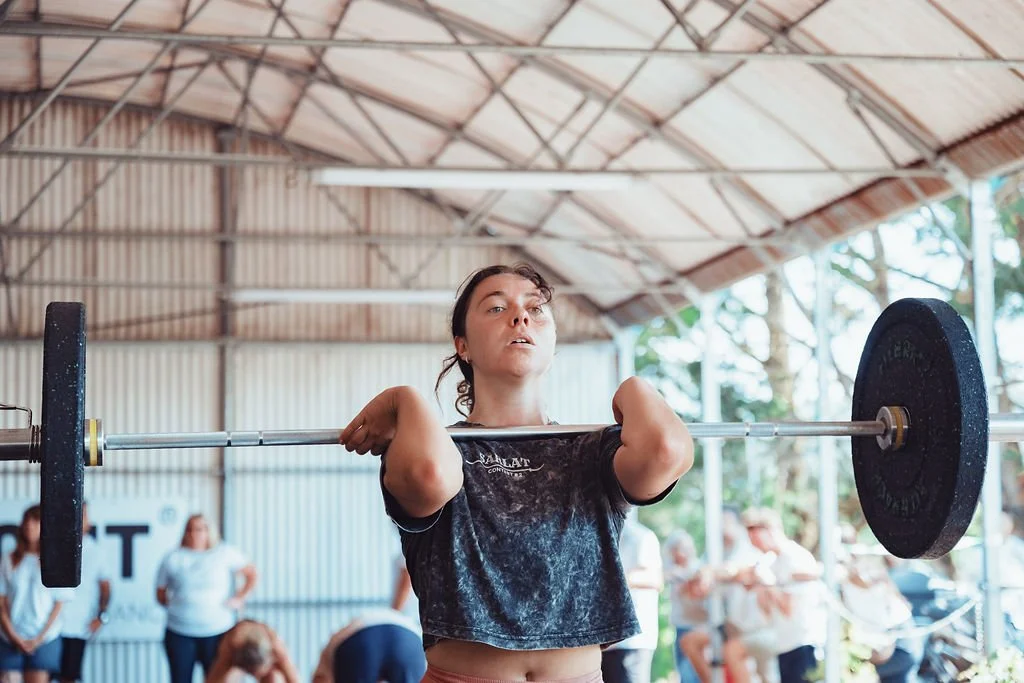 Une femme soulève une barre de musculation lors d'une séance d'entraînement dans une salle de sport, avec d'autres personnes en arrière-plan