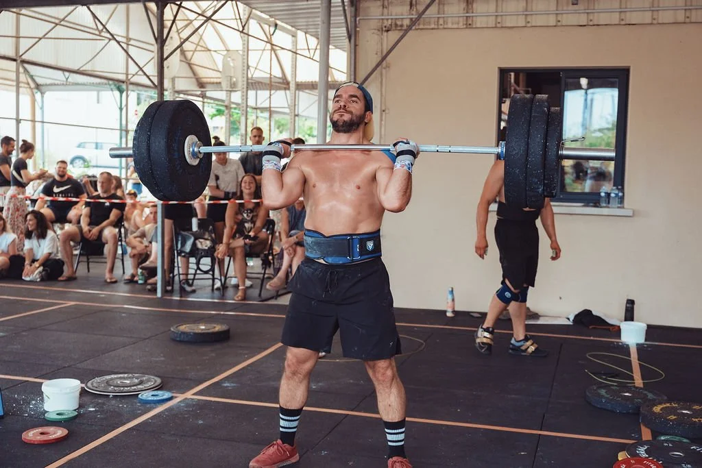 Un homme en train de soulever une barre de poids lors d'une compétition de musculation, avec un public assis en arrière-plan.