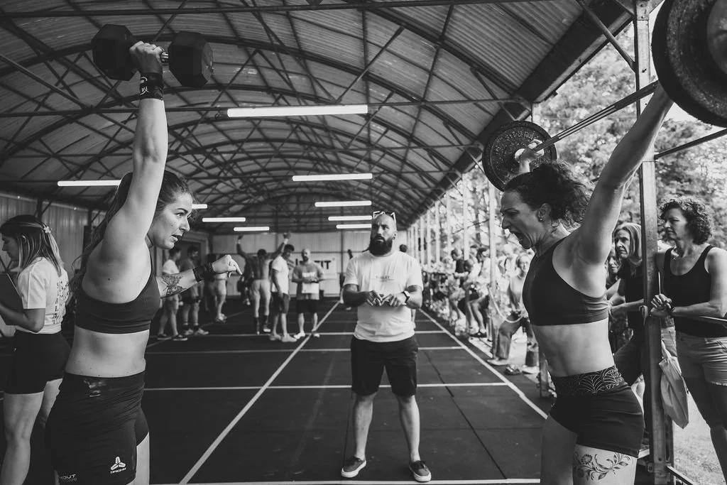 Deux jeunes femmes s'entraînent au CrossFit avec des haltères sous une tente, entourées d'autres personnes et d'un entraîneur observant