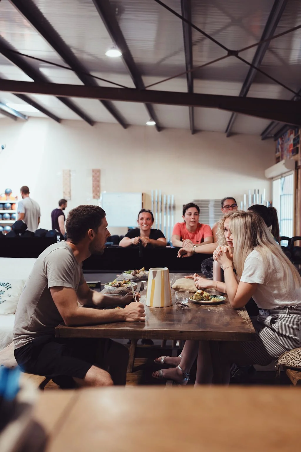 Groupe de jeunes adultes assis autour d'une table en bois en train de discuter, avec des plats de nourriture devant eux, dans un espace intérieur lumineux avec un plafond en métal et des murs clairs.