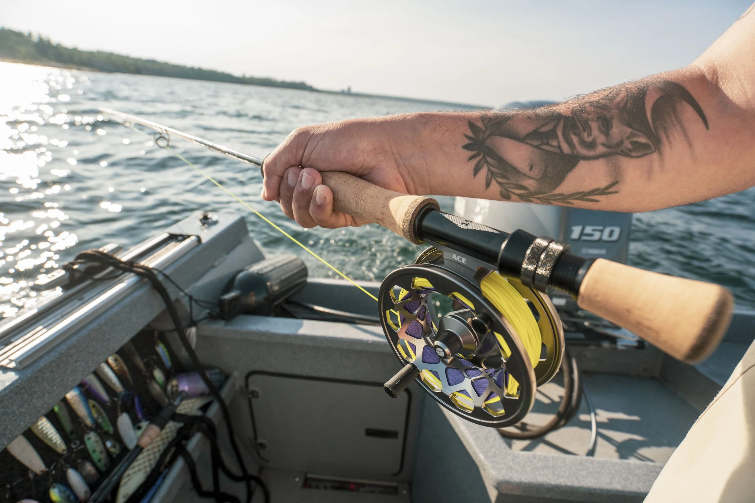 A person fishing on a boat with a fishing rod and reel, wearing a tattooed arm, on a calm body of water with the shoreline and trees in the background.