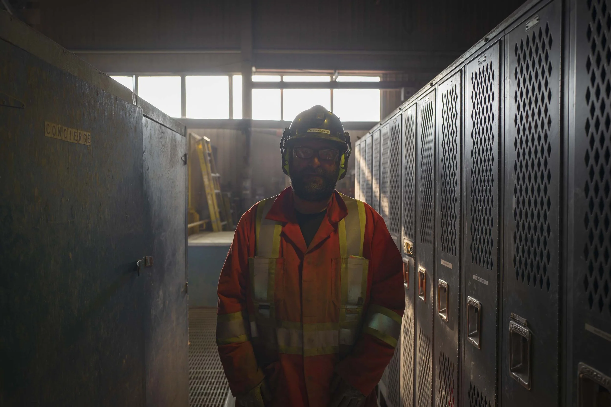 A worker in safety gear in an industrial setting with lockers and equipment in the background.