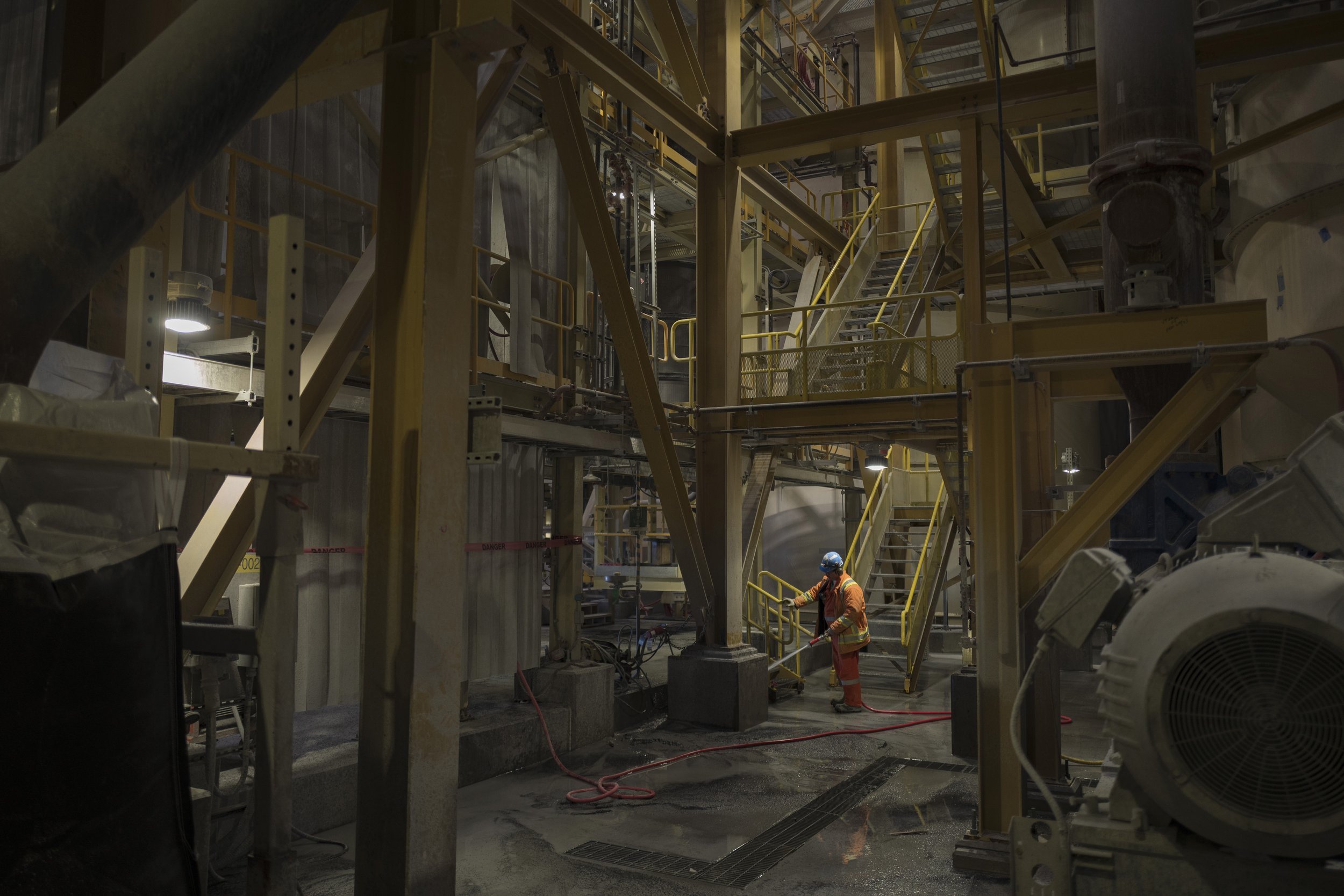 Worker wearing safety gear, including a blue helmet and orange suit, cleaning or inspecting the floor inside an industrial facility with yellow stairs and metal structures.