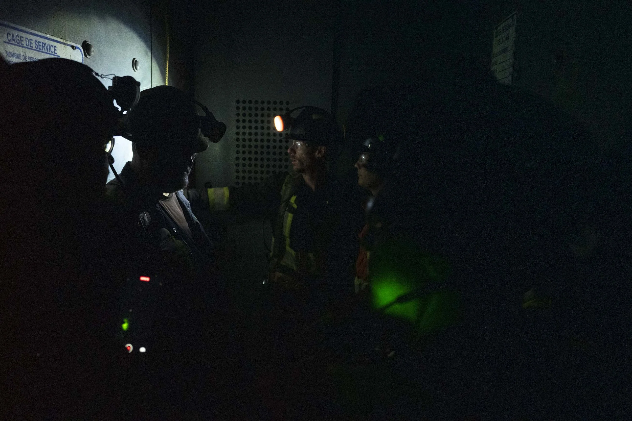 Group of four emergency responders in a dark room, wearing helmets with headlamps, illuminated by green and red lights, standing near a wall with a service cage sign.