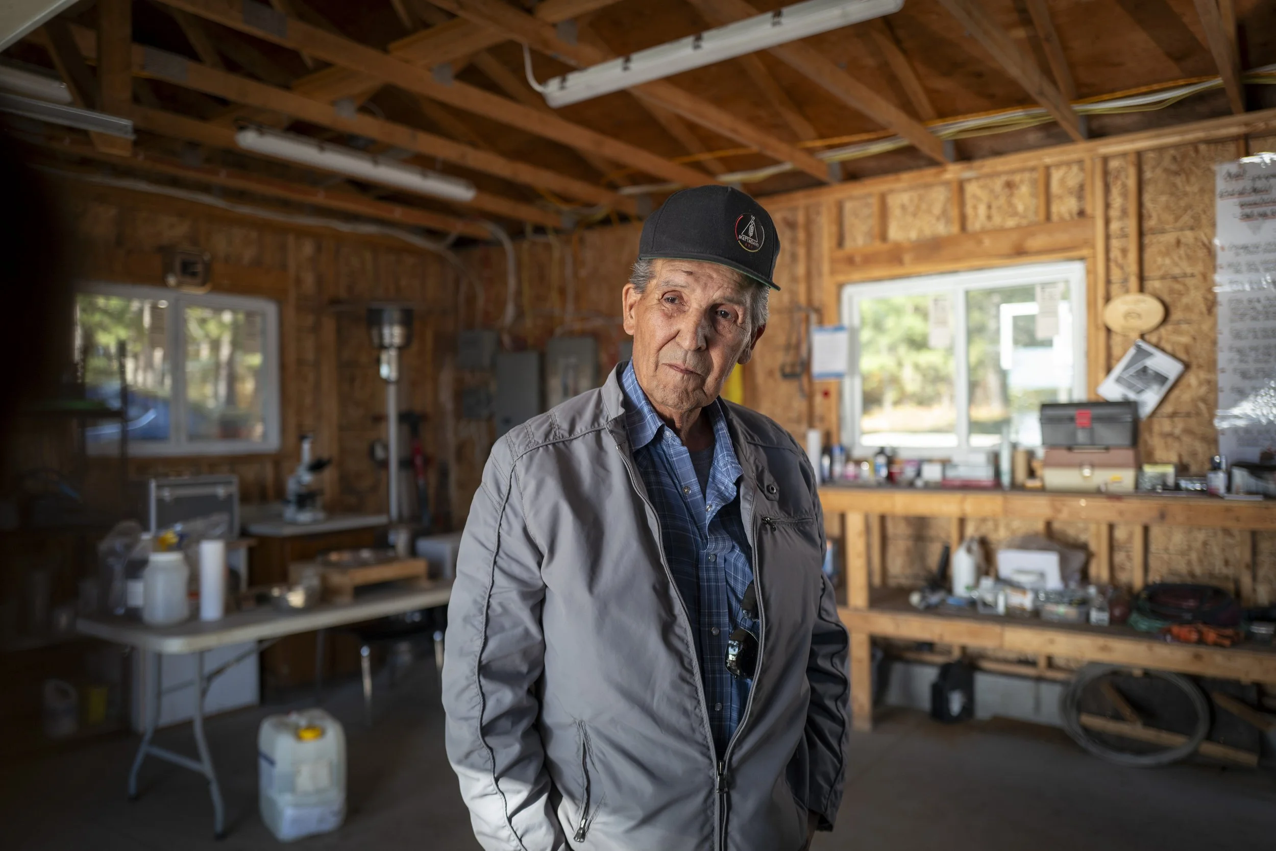 An elderly man with a serious expression, wearing a gray jacket and a black baseball cap, standing inside a wooden workshop with tools and supplies on shelves and a window showing greenery outside.