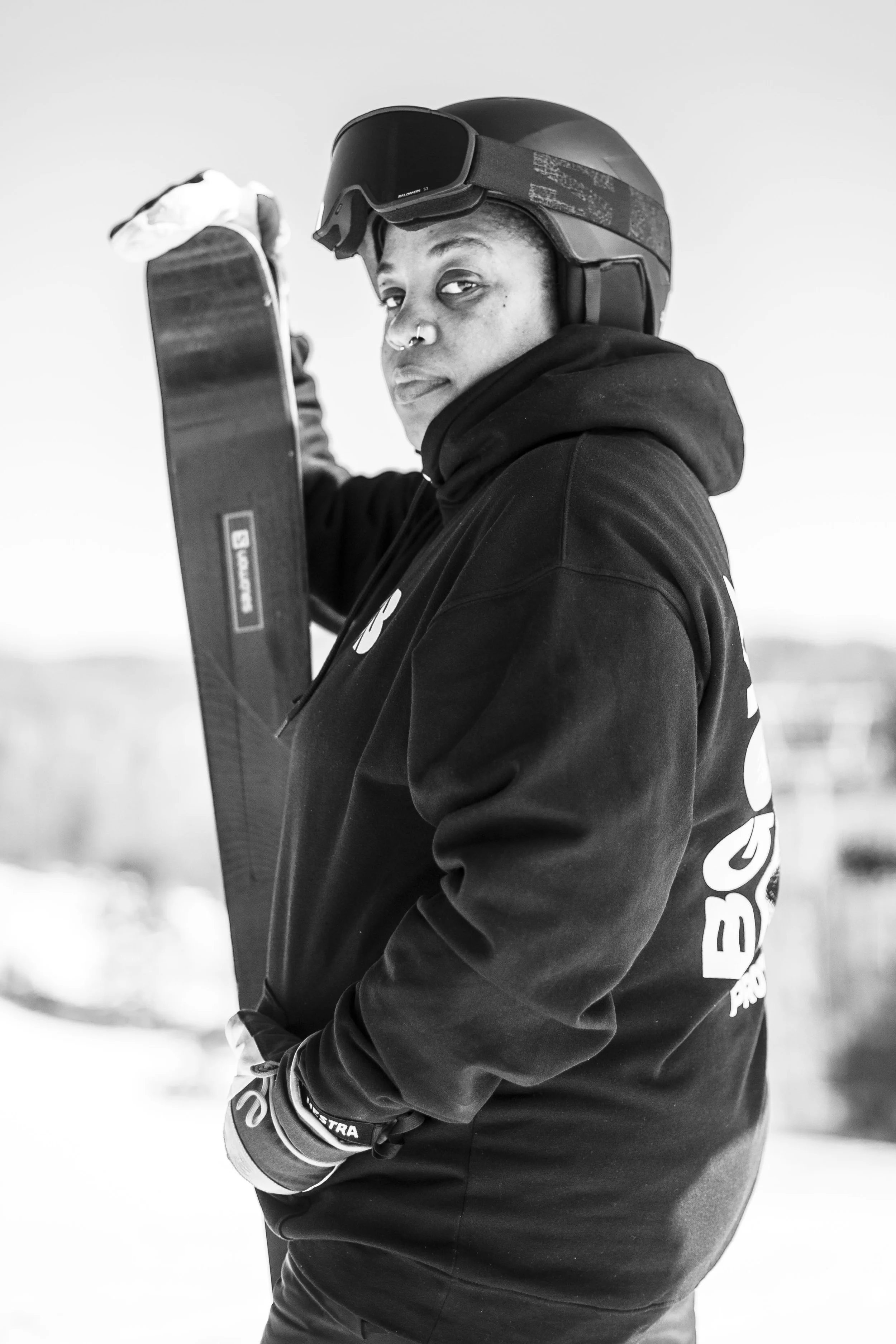 Black and white photo of a person wearing a helmet, hoodie, and ski gloves holding a ski pole, outdoors in a snowy environment.