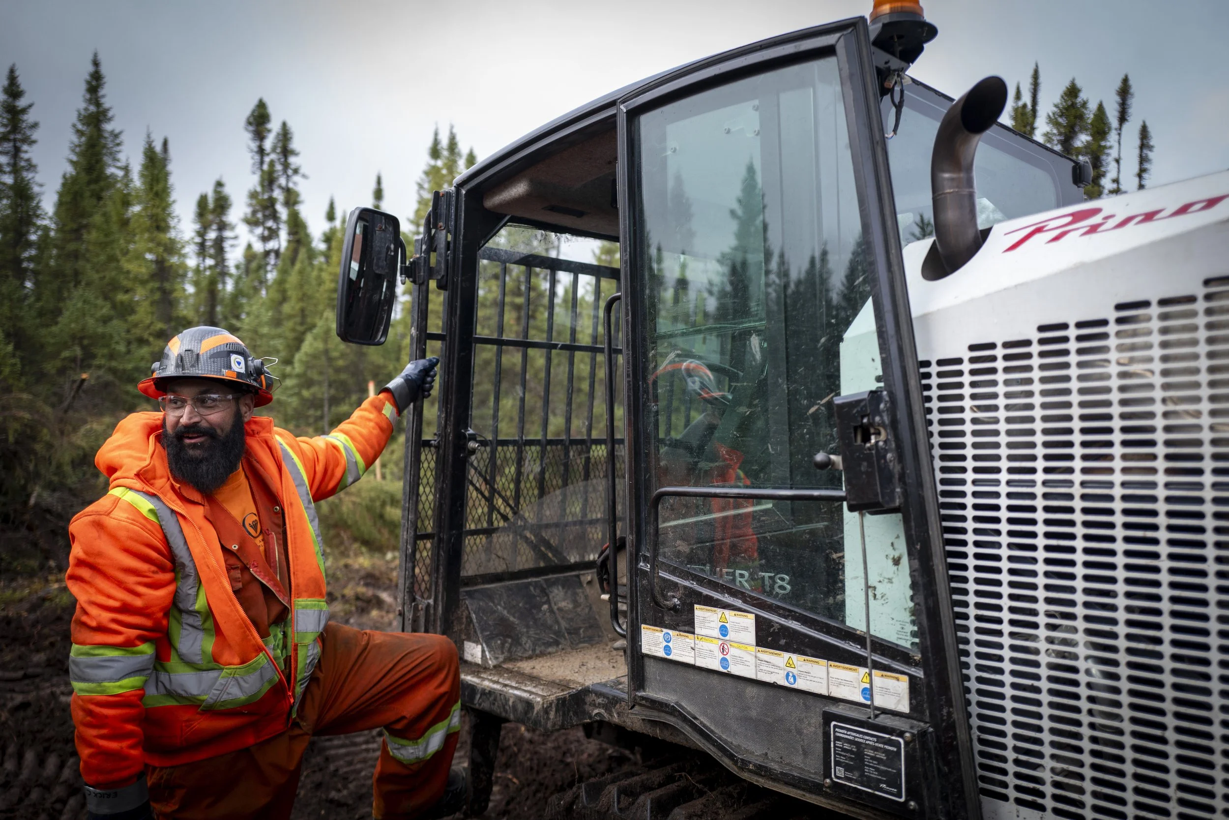 A construction worker wearing an orange safety jacket and helmet, standing next to a heavy construction vehicle in a forested area.