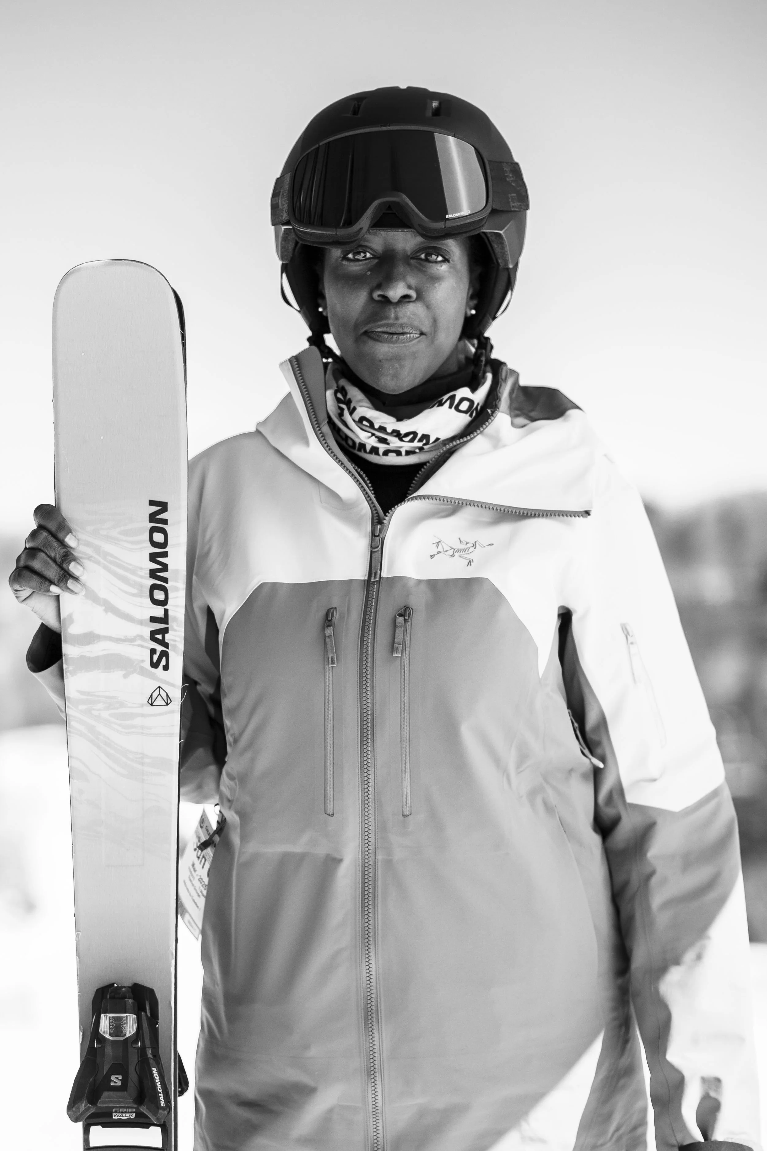 A woman dressed in skiing gear, holding a ski pole, wearing a helmet and goggles, standing outdoors with a snowy background.